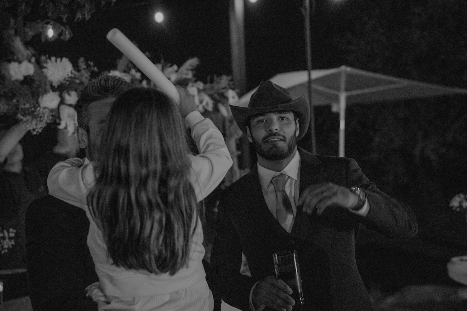 Groom in cowboy hat dances with guest at lively outdoor wedding reception in black and white photo.