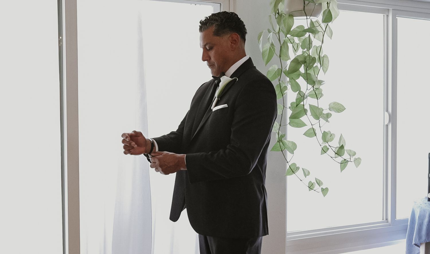 Elegant groom in black tuxedo adjusting cufflinks near bright window with green ivy plant.