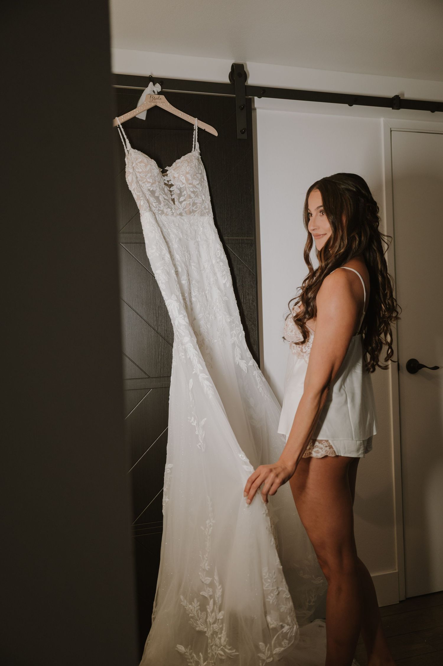 Bride in lingerie admiring her white lace wedding gown hanging on a barn door in a softly lit room.