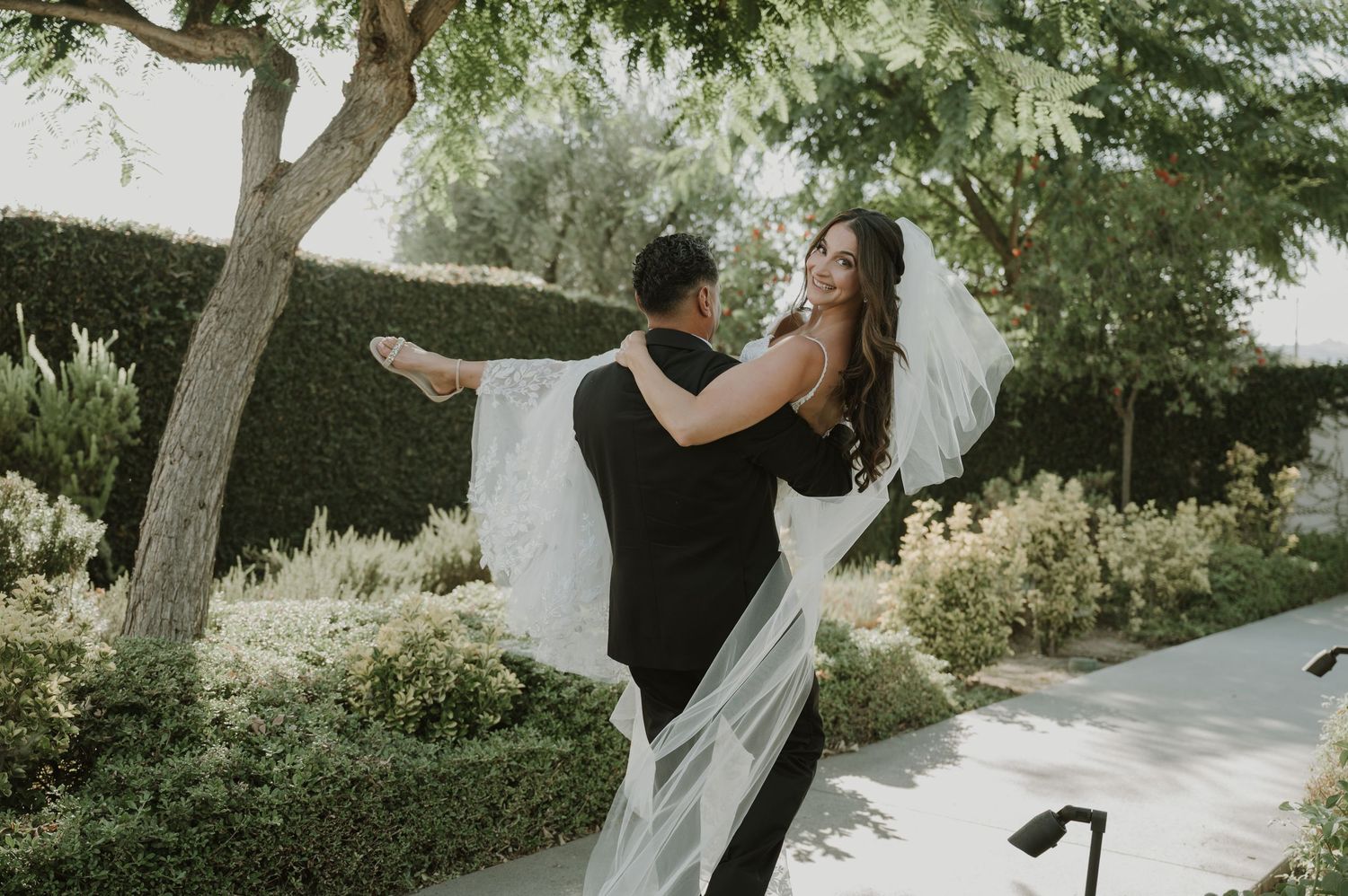 Groom lifting bride in wedding dress with flowing veil outdoors among lush green hedges and trees.