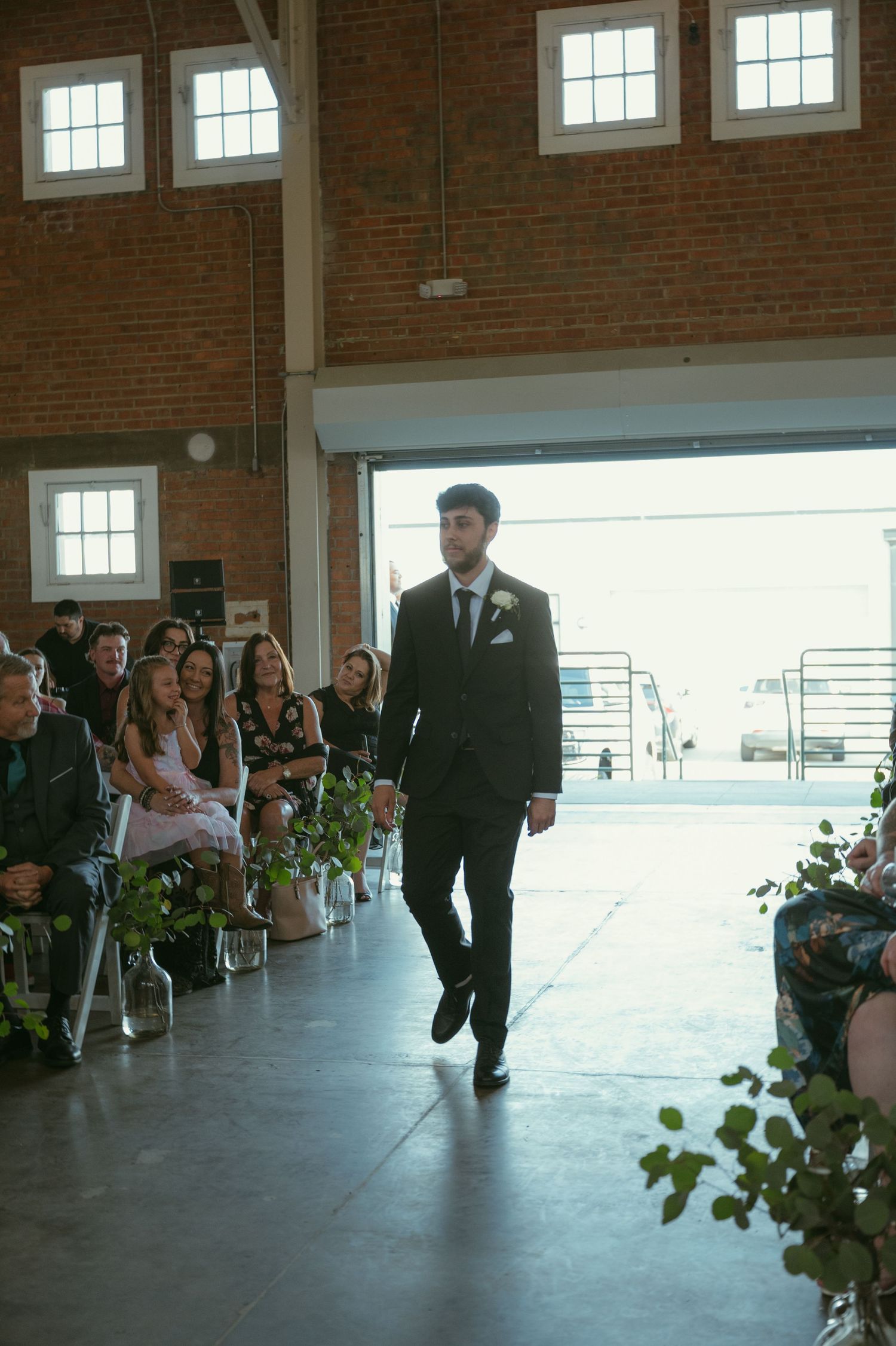 Groom walks down the aisle in a black suit at an industrial brick wedding venue.