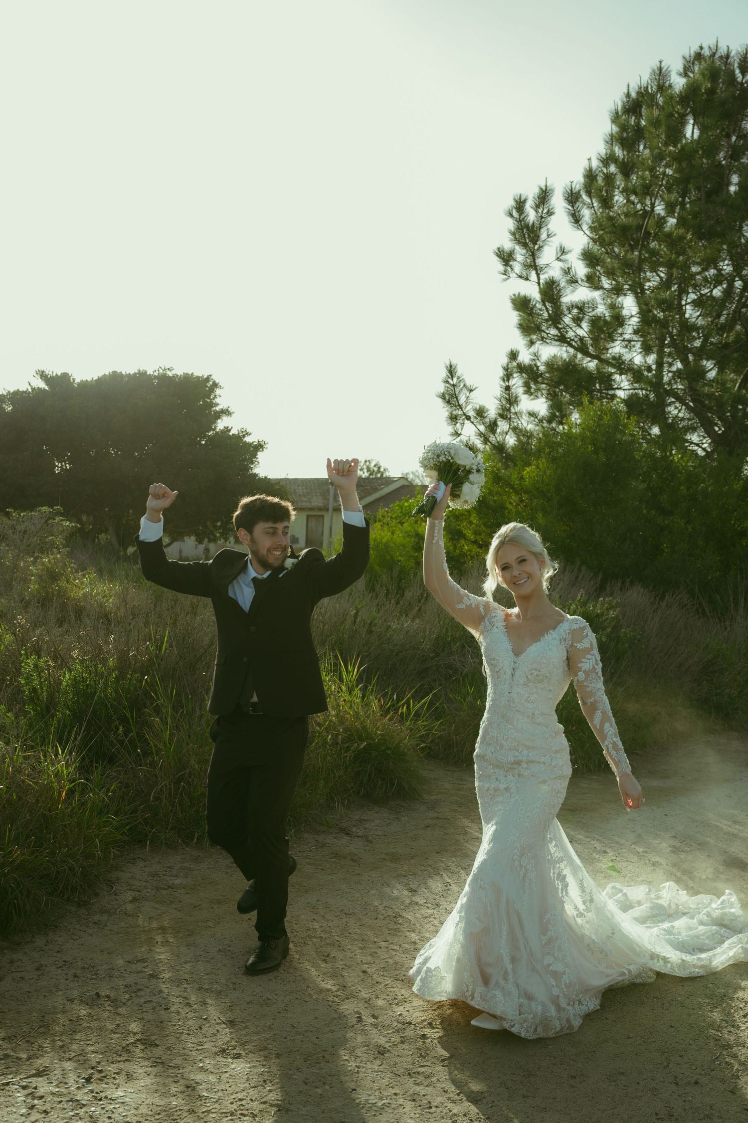 Joyful newlyweds celebrate on a sunlit dirt path, bride in lace gown holding bouquet, groom in black suit, arms raised.