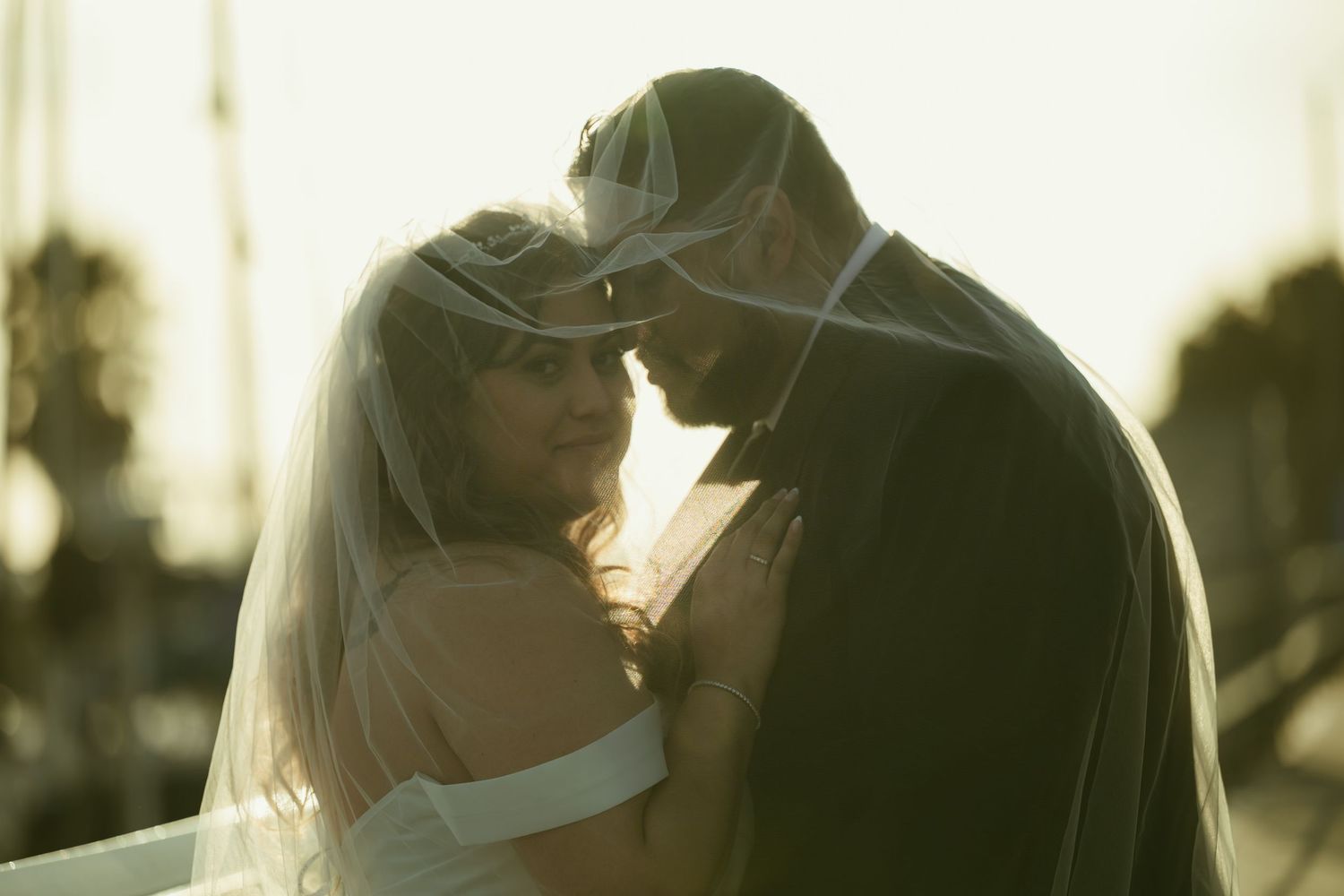 A bride and groom share an intimate moment under a veil at sunset near a marina on their wedding day.