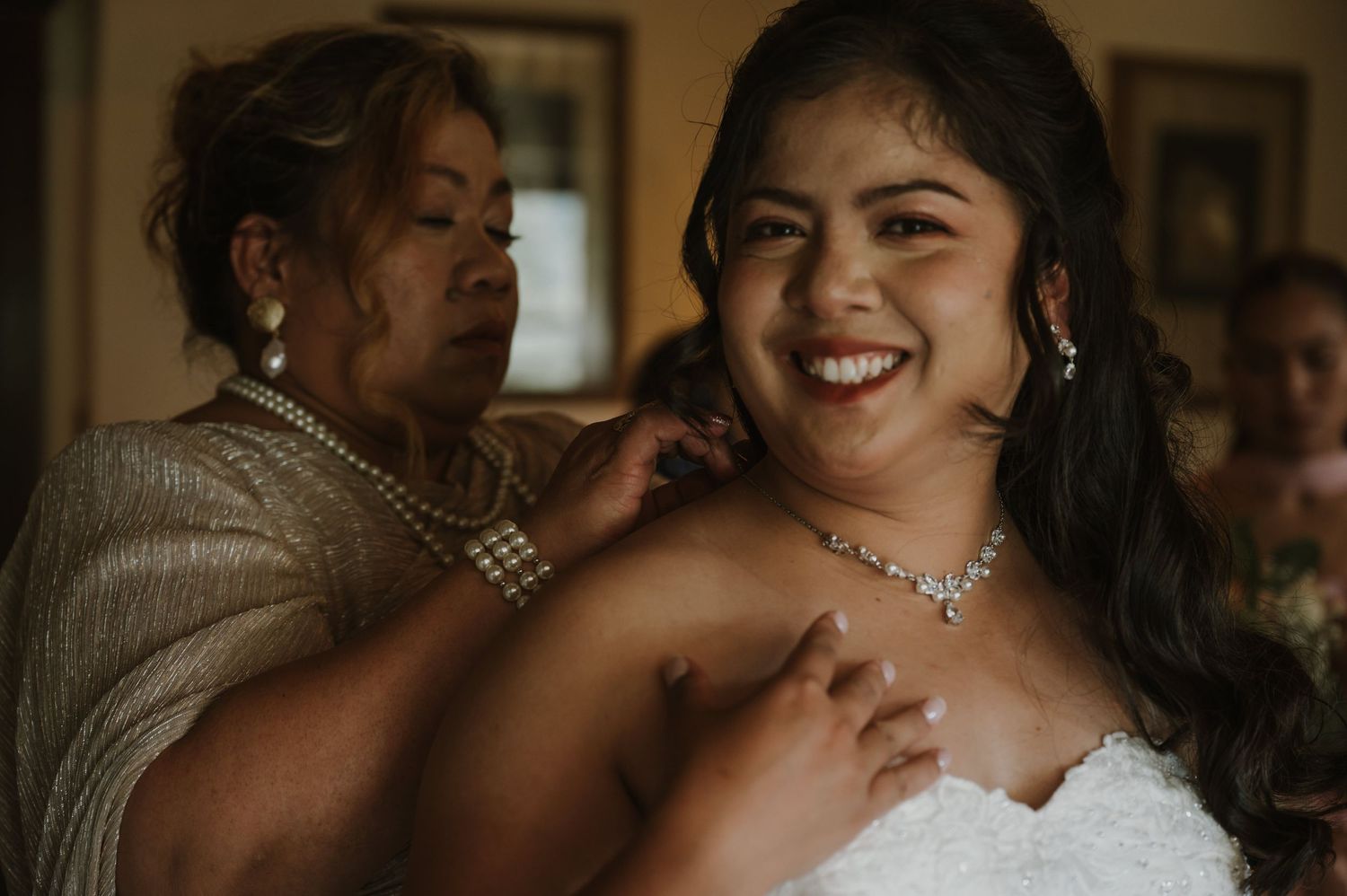 A smiling bride in a strapless gown gets her pearl necklace fastened by an older woman in a beaded dress.