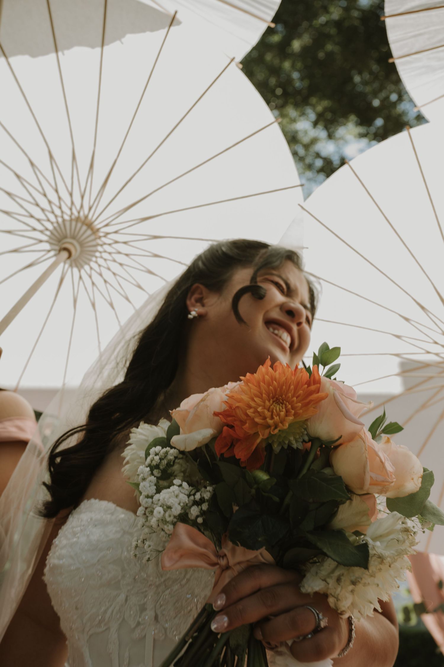 Smiling bride in white dress holds peach and orange bouquet under elegant white parasols outdoors.