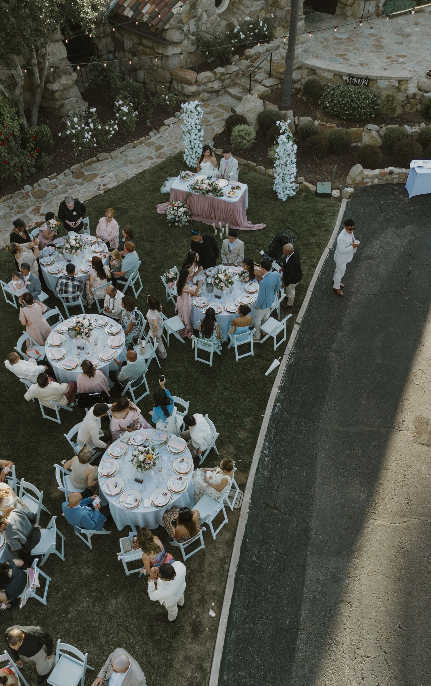 Aerial view of an elegant outdoor wedding reception with round tables, white chairs, and floral centerpieces on a green lawn.