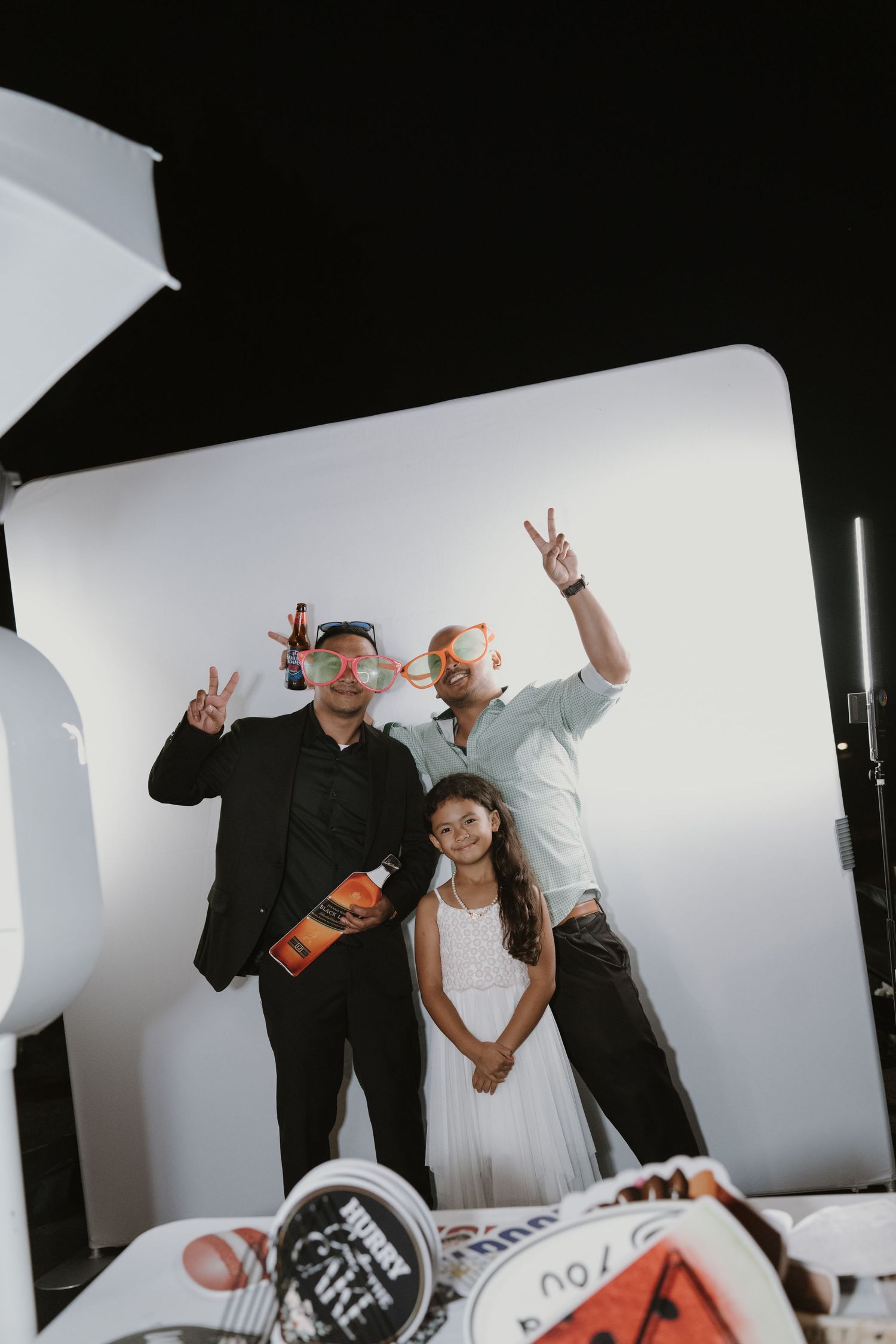 Two men and a young girl pose playfully in a wedding photo booth with fun props and peace signs.