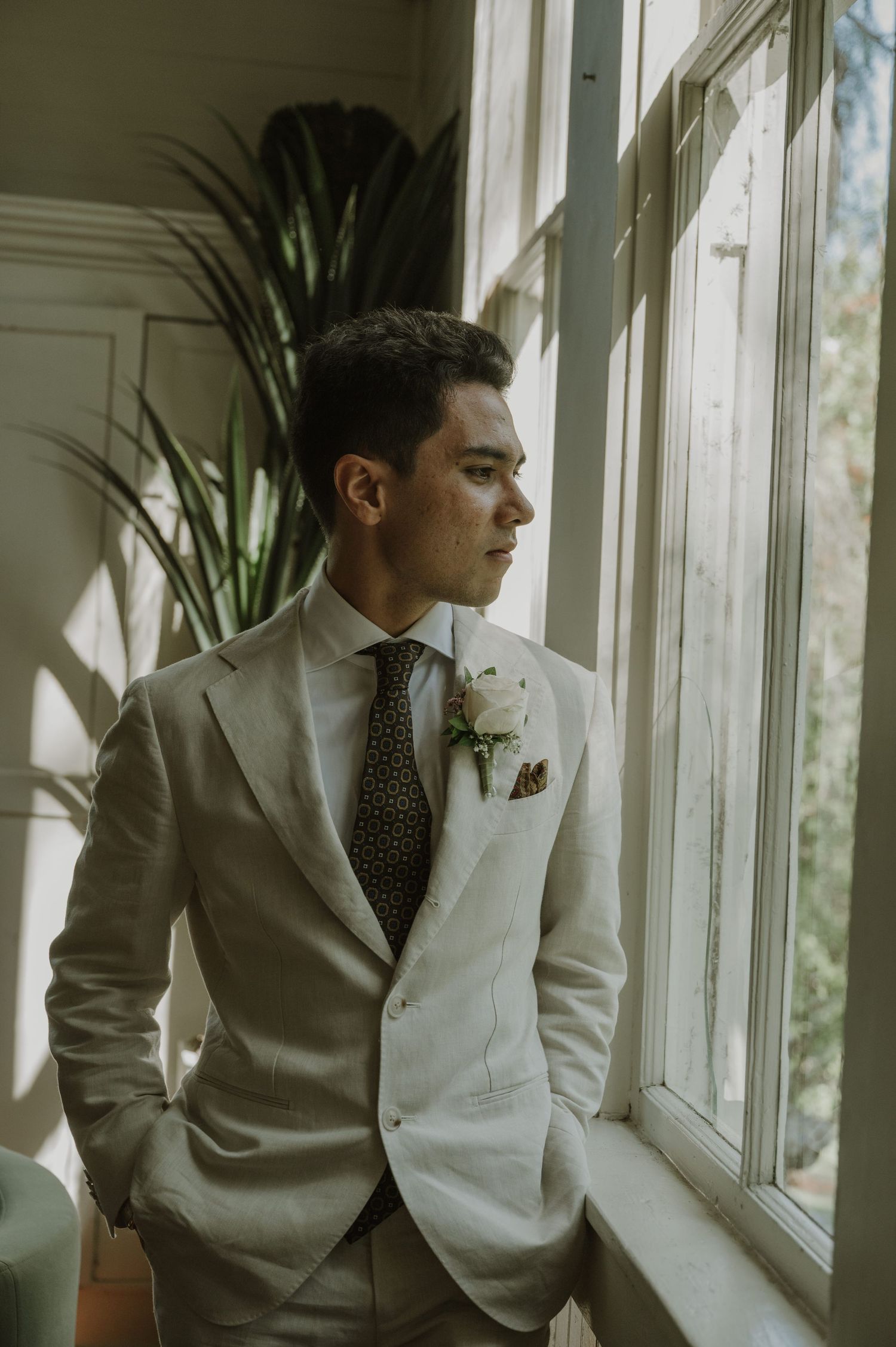 Dapper groom in cream linen suit with boutonniere gazes out sunlit window on wedding day.