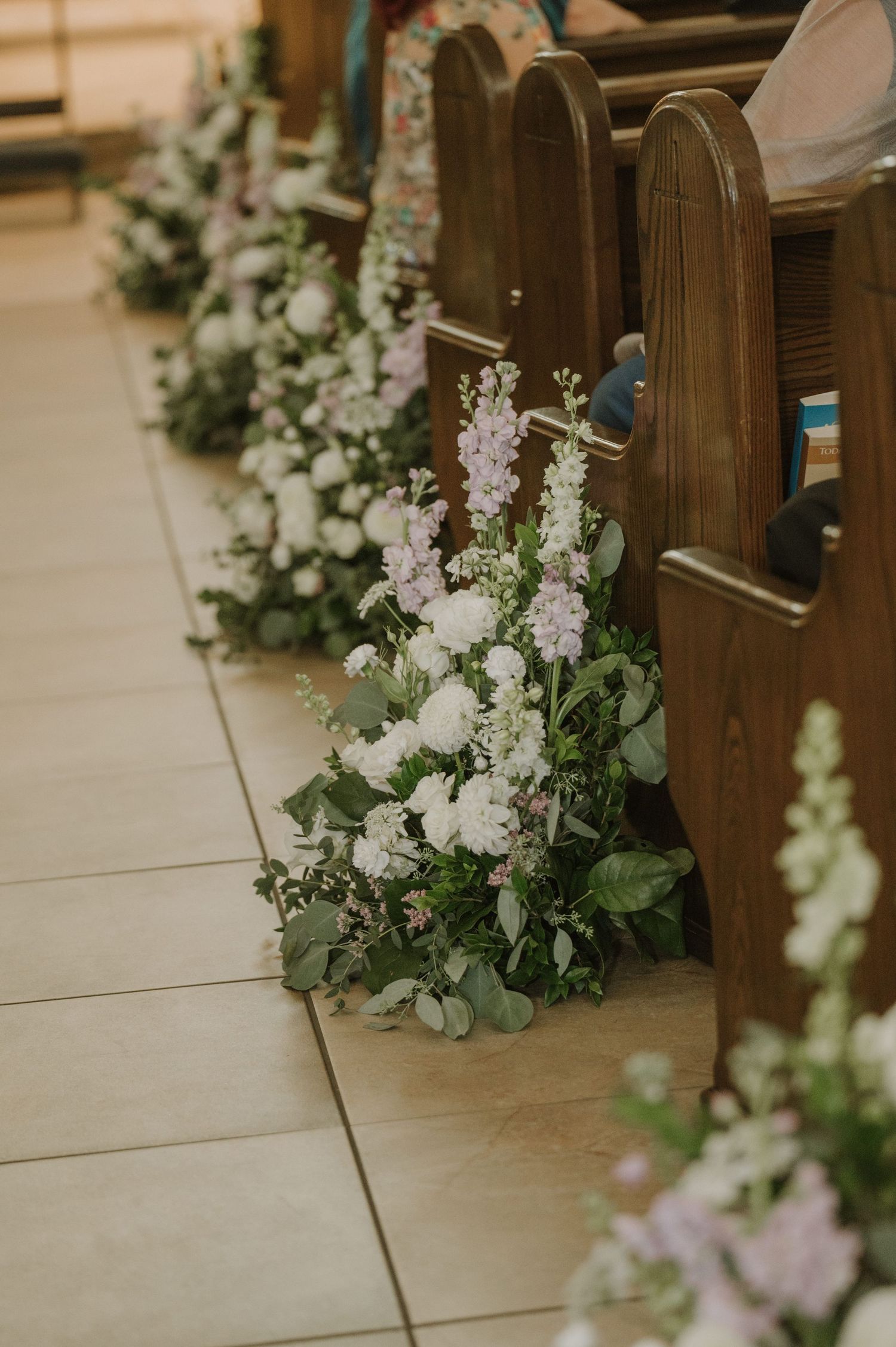 White and lavender floral arrangements line wooden church pews along a tiled aisle for a wedding ceremony.