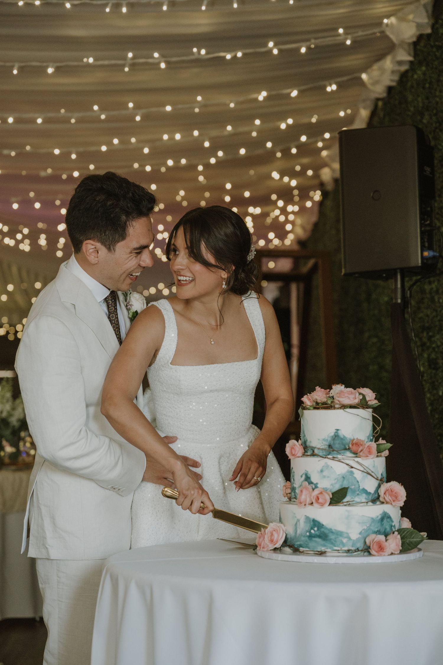 Bride and groom cut three-tier wedding cake decorated with pink roses and teal accents under fairy lights.