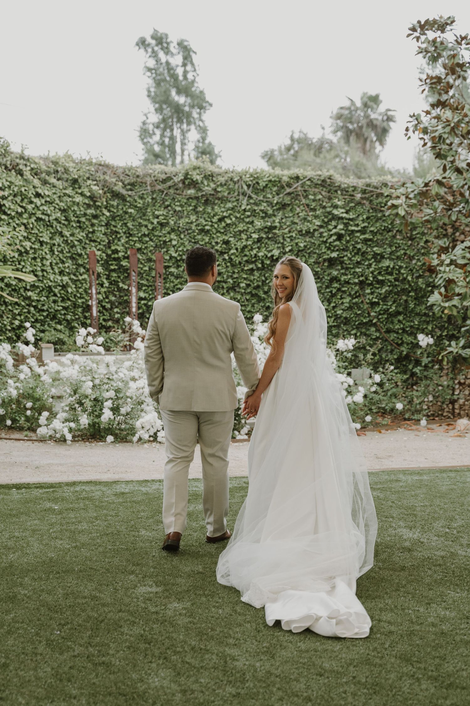 Bride in white gown and veil holds grooms hand walking through lush garden wedding venue.