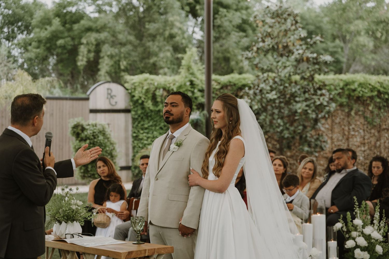 Bride and groom stand at outdoor wedding altar as officiant speaks during garden ceremony.