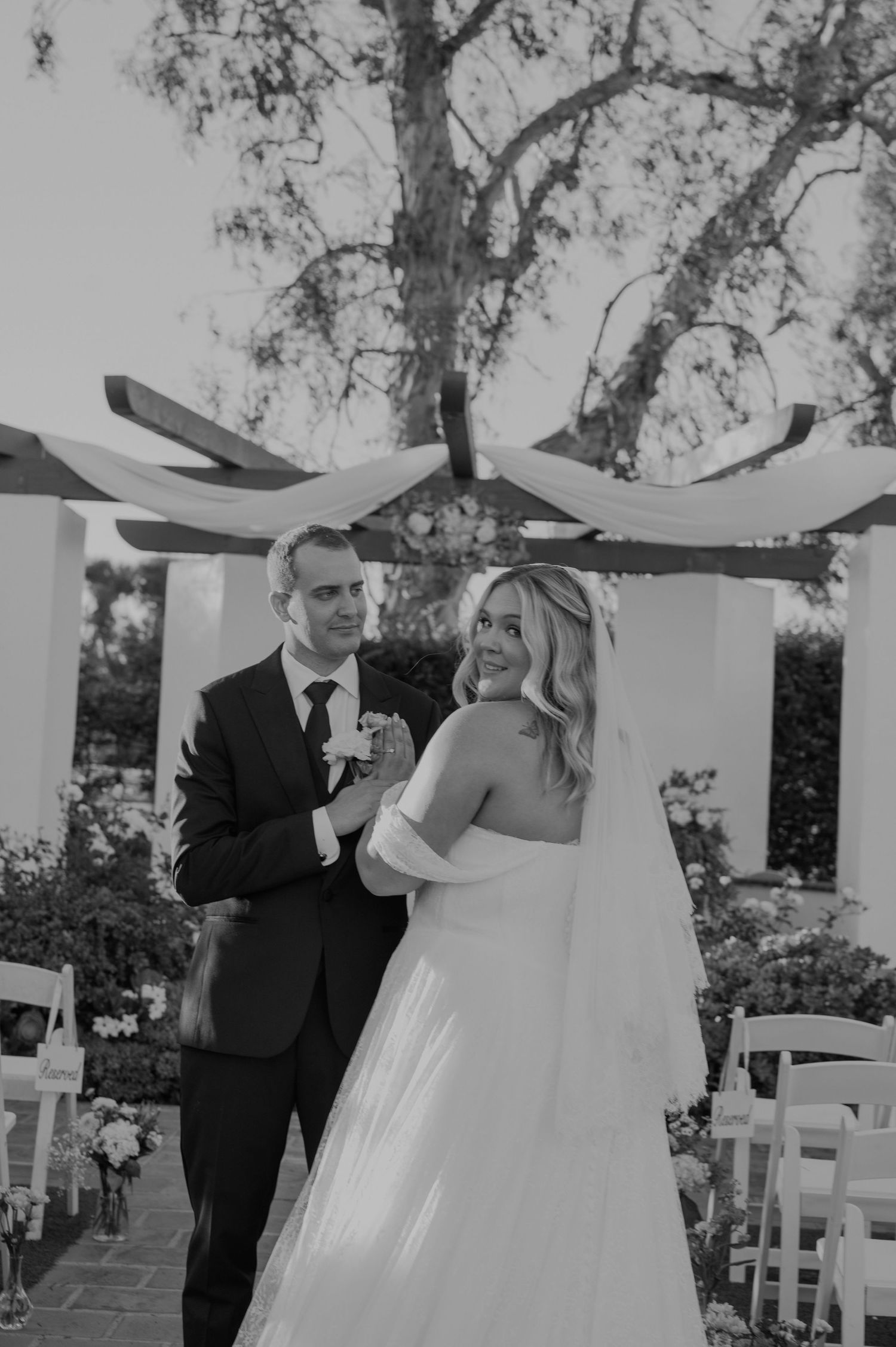 Black and white photo of a smiling bride in a white gown holding hands with her groom in a suit at an outdoor wedding.