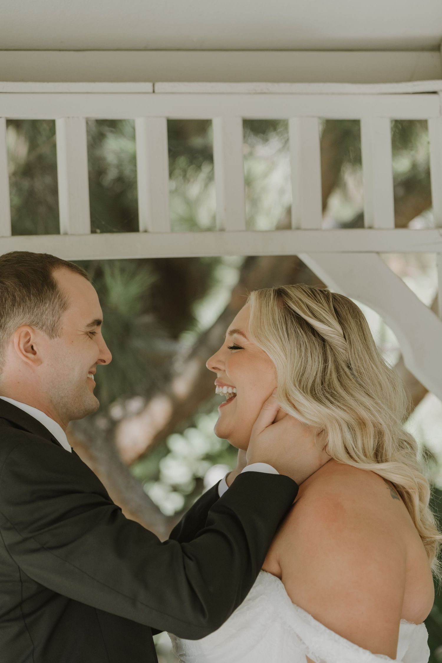 A groom tenderly holds his laughing bride's face during a romantic wedding moment outdoors.