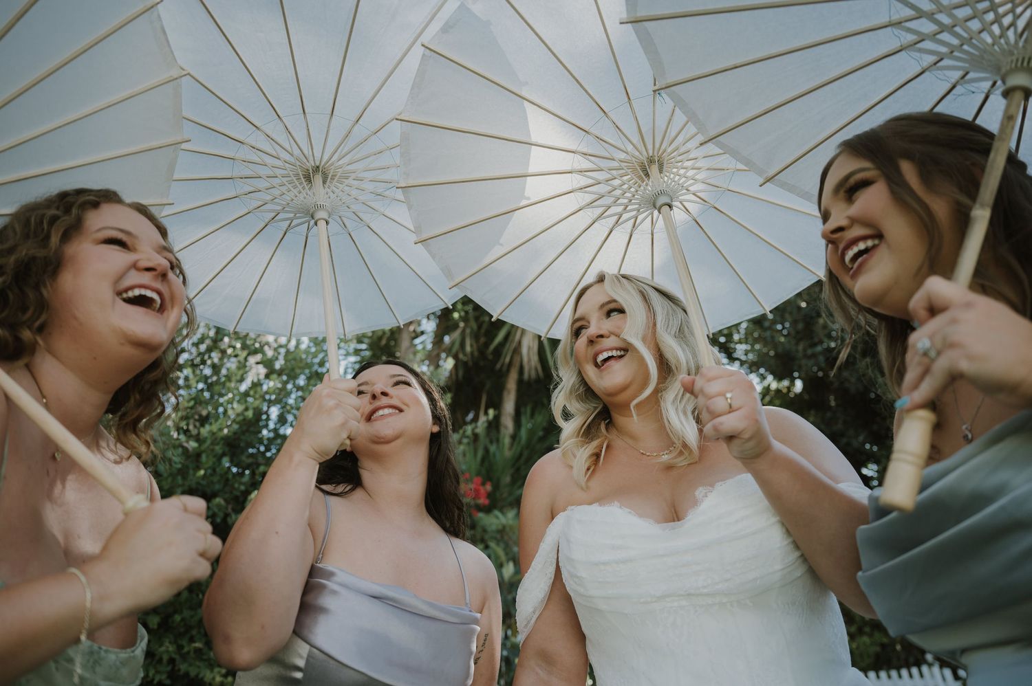 Bride and bridesmaids laughing together under white parasols at an outdoor wedding celebration.