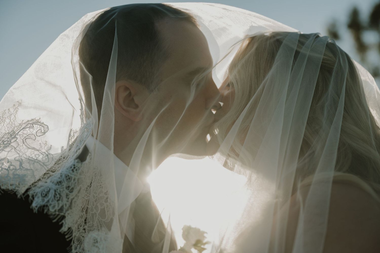 Bride and groom sharing a romantic kiss under a flowing wedding veil with soft backlit sunlight.