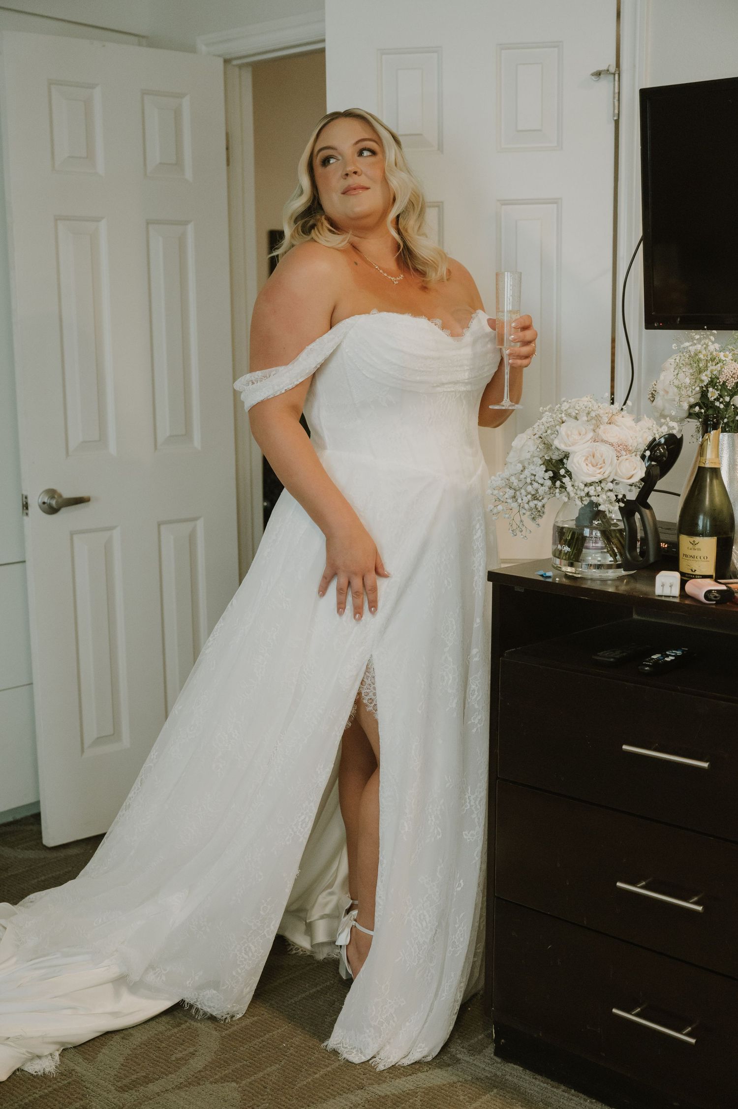 Bride in off-shoulder white wedding dress with slit holding champagne glass before ceremony.