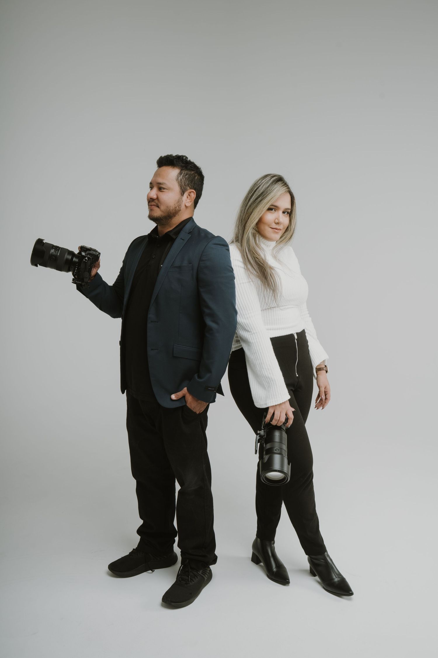 A male and female photographer pose back-to-back holding cameras in a professional studio setting.