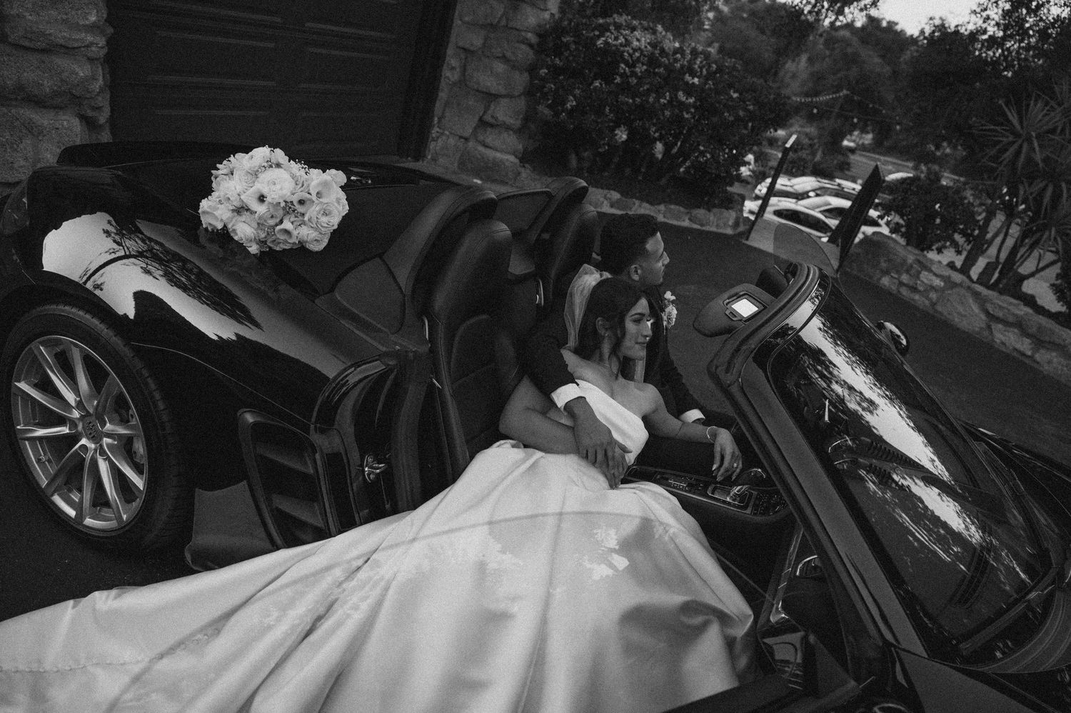 Black and white photo of a bride in a flowing wedding dress getting into a luxury car at an outdoor venue.