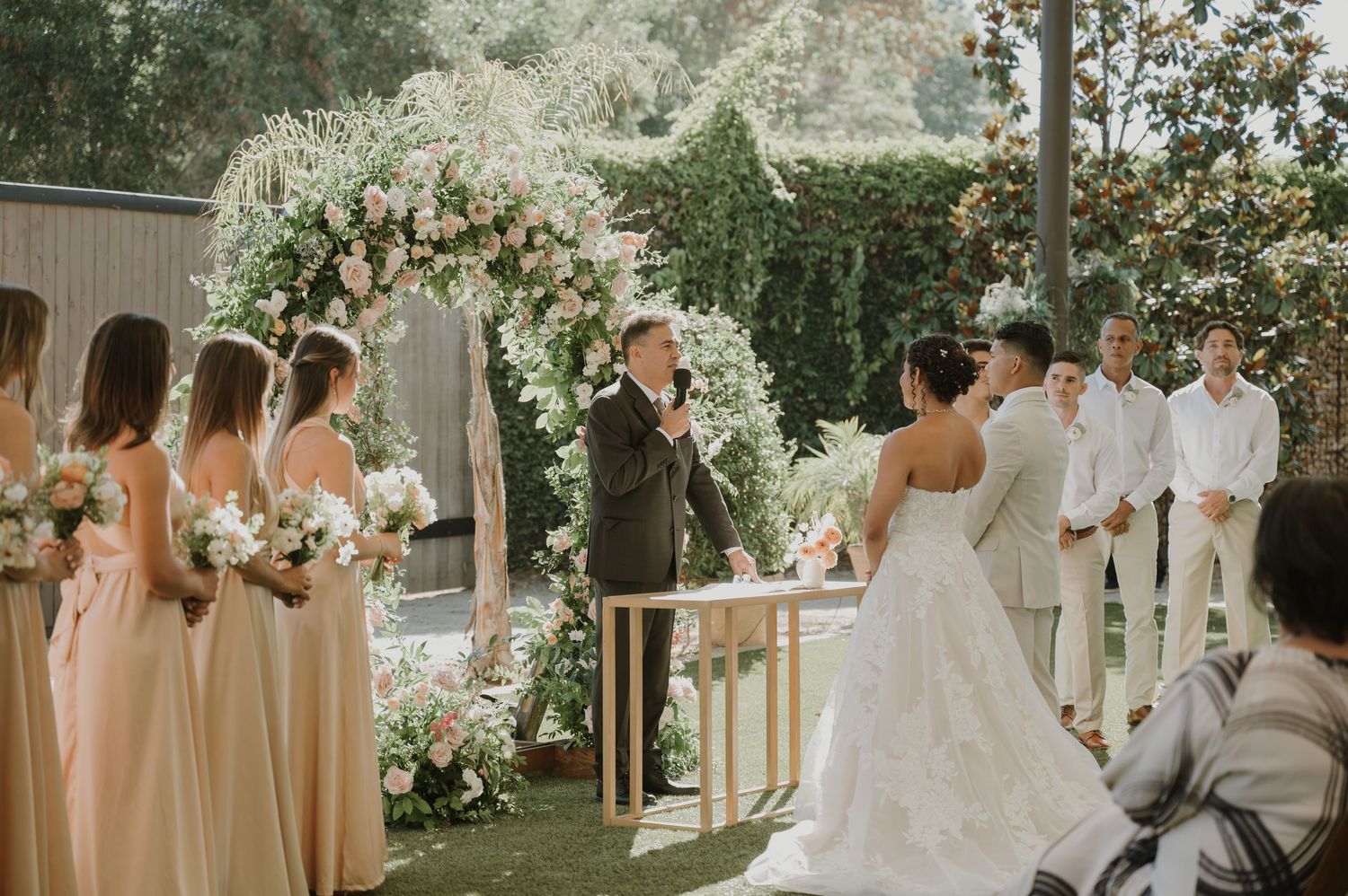 Outdoor wedding ceremony with bridesmaids in blush dresses, floral arch, and guests seated on a green lawn.