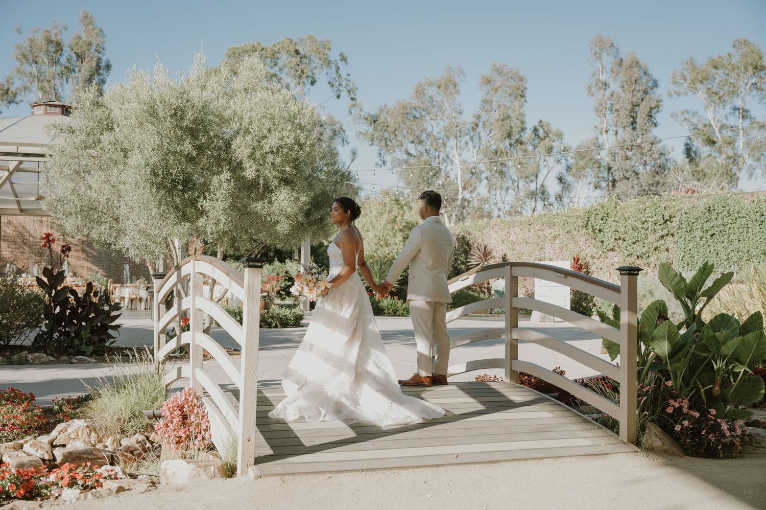 Bride and groom stand on a white wooden bridge surrounded by lush gardens on their wedding day.