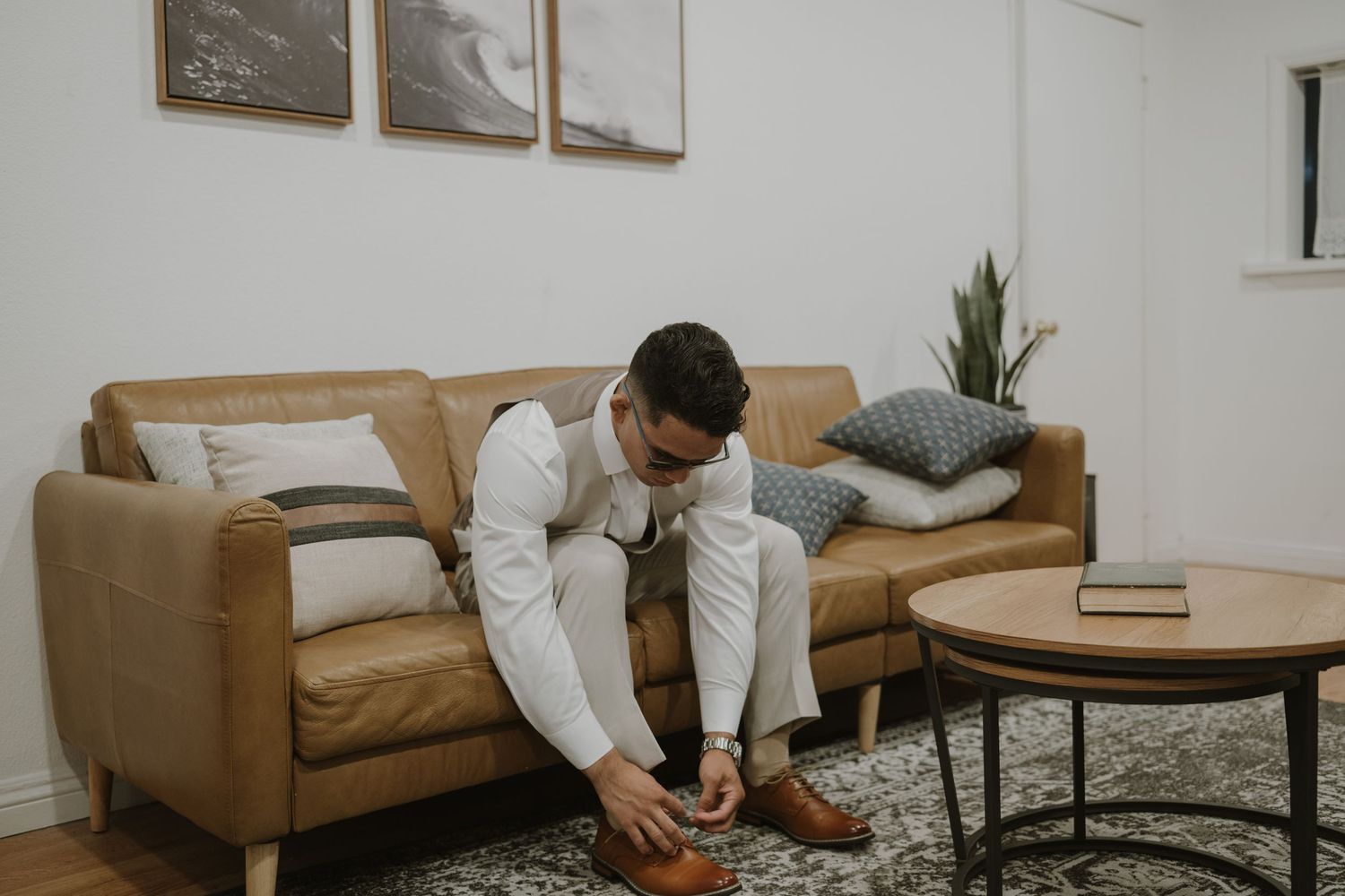 Groom in white shirt and trousers ties his brown dress shoes on a leather sofa.