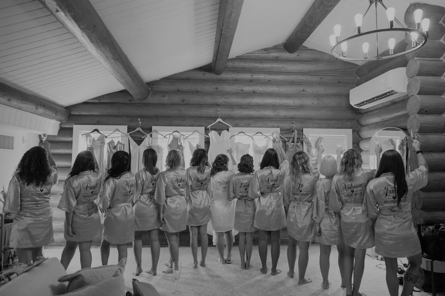Bridesmaids in matching robes standing with backs to camera in a rustic log cabin room, black and white.