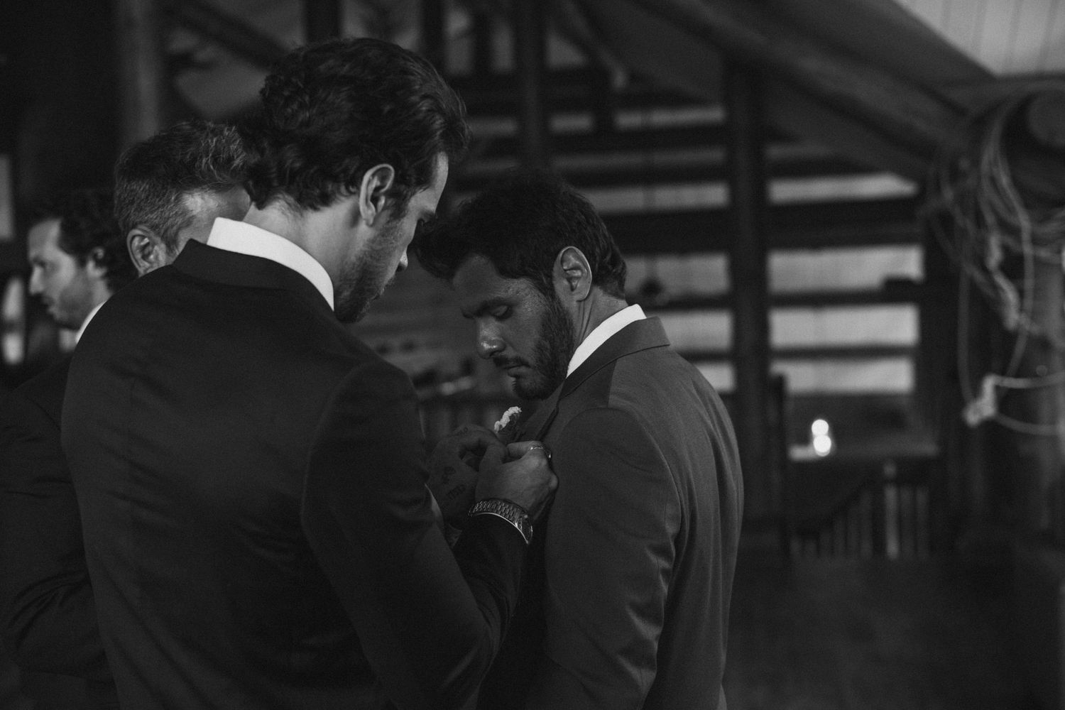 Groomsmen in suits helping pin boutonnieres in a dimly lit rustic barn venue.