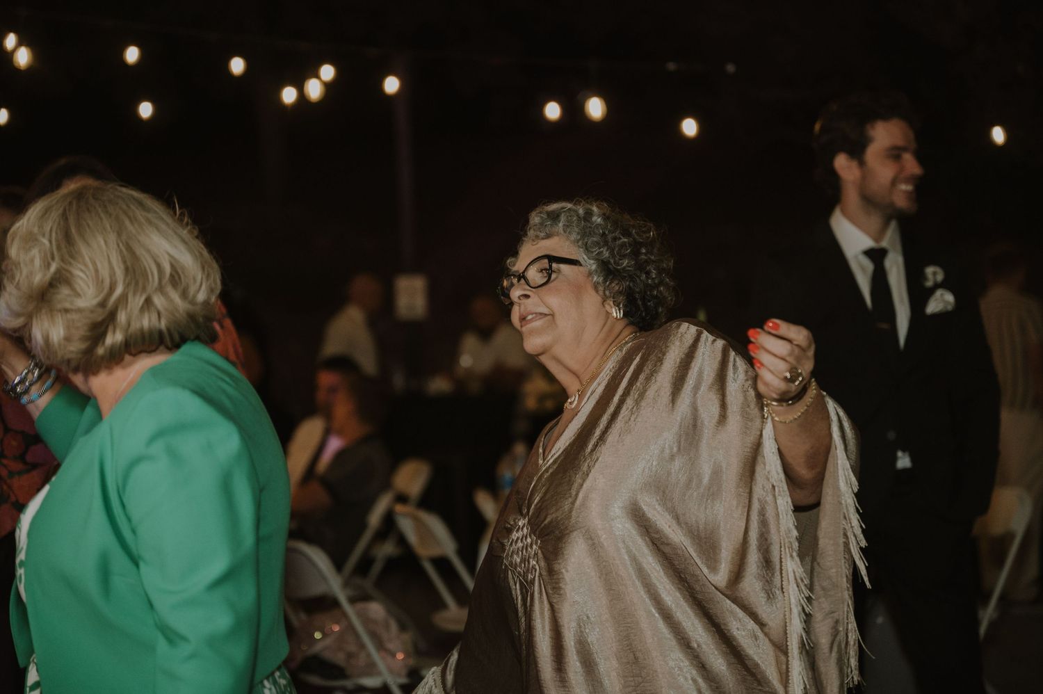 Elderly woman in a gold shawl smiling and dancing joyfully at an outdoor wedding reception with string lights.