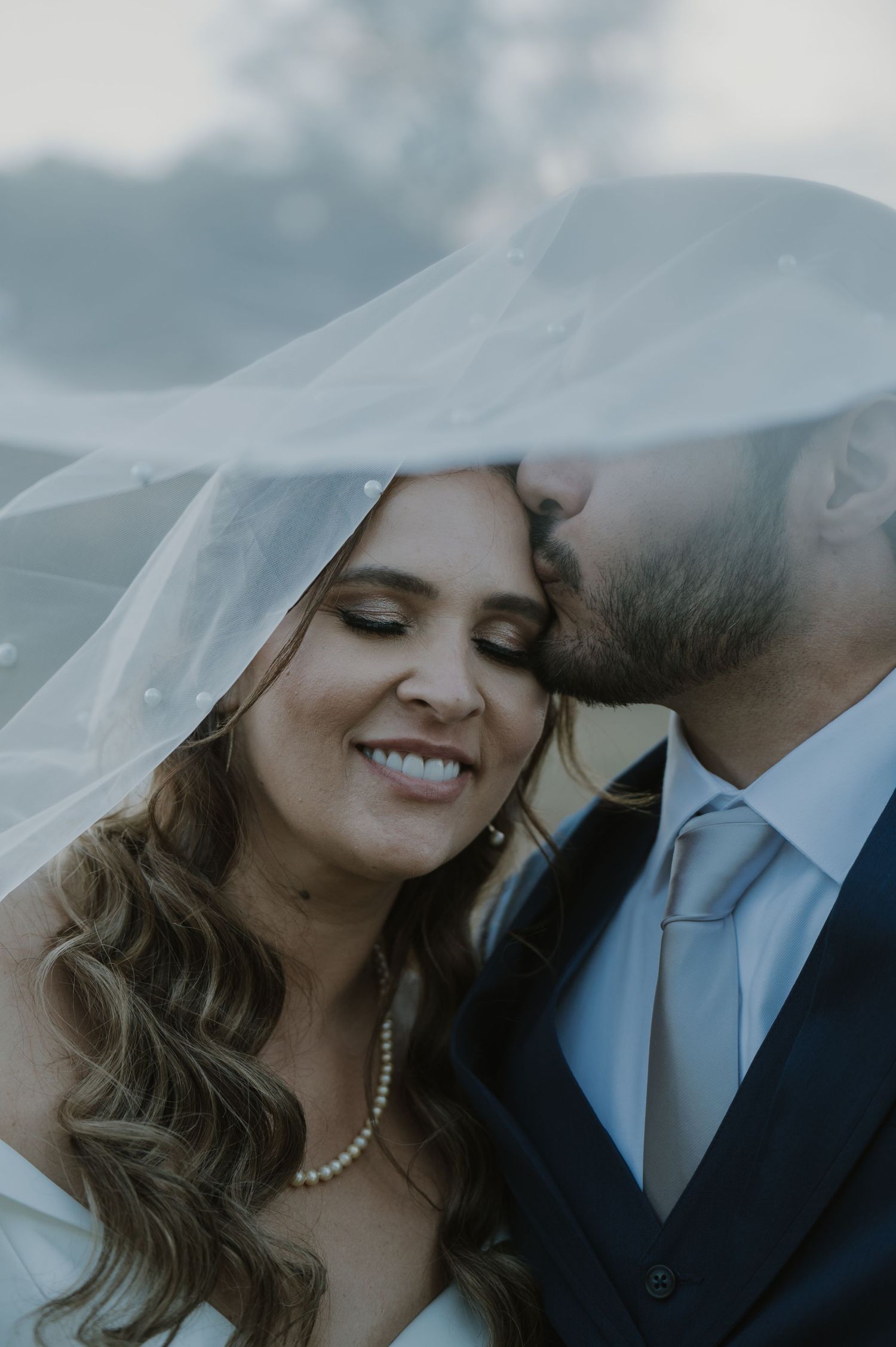 Bride smiles as groom kisses her cheek under flowing veil, wearing pearl necklace and elegant bridal gown.