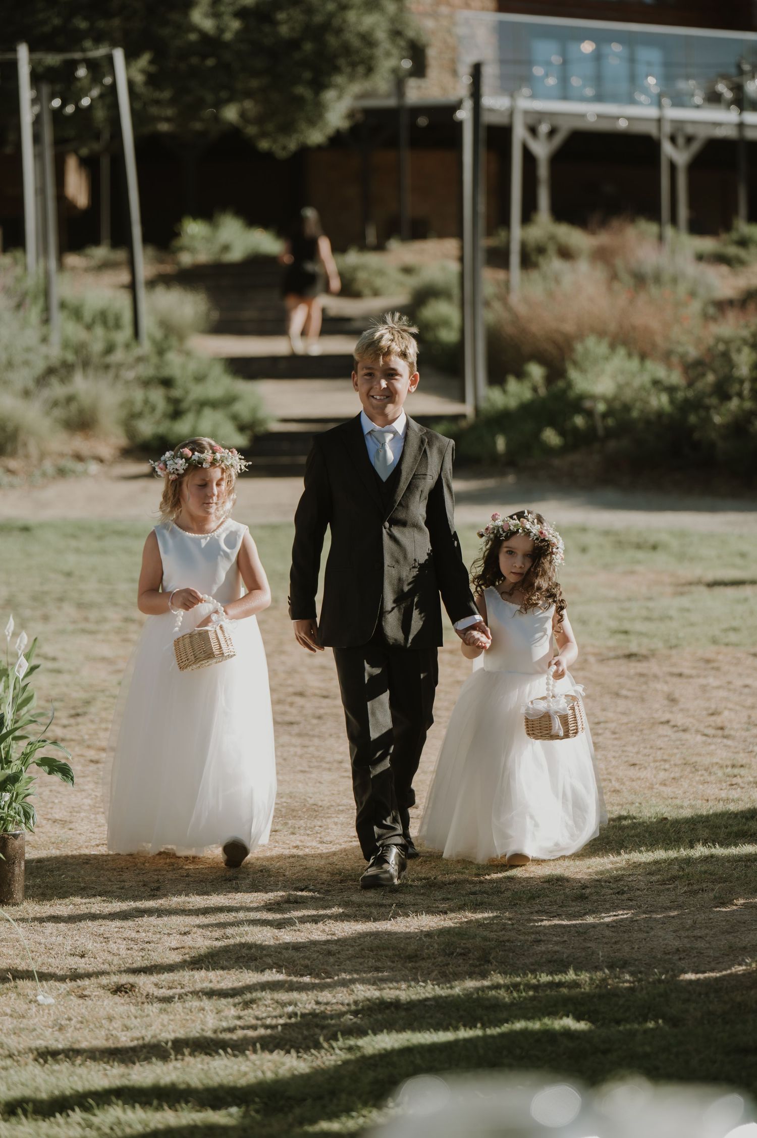 A young boy in a suit escorts two flower girls in white dresses and floral crowns down the aisle at an outdoor wedding.