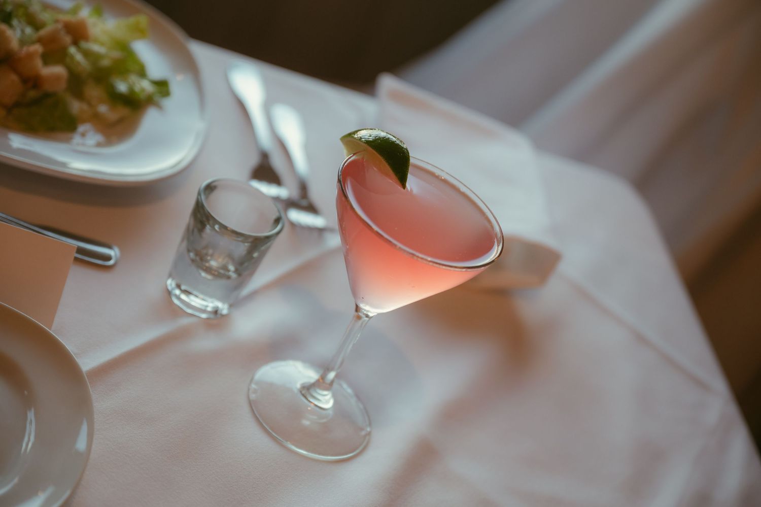 Pink cosmopolitan cocktail with lime garnish in a martini glass on a white tablecloth at a restaurant.