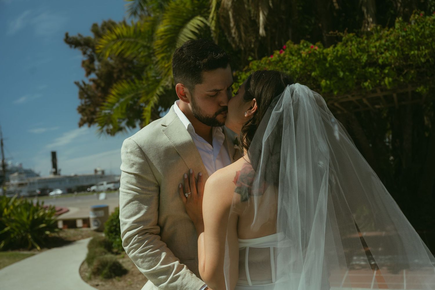 A couple shares a romantic moment in wedding attire, bride in veil, groom in tan suit, outdoors under bright sunlight.
