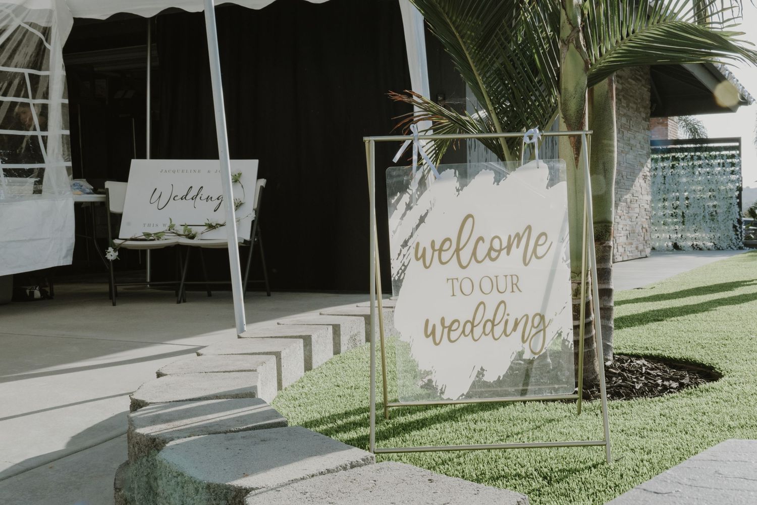Elegant outdoor wedding welcome sign on gold frame stand with white acrylic board and green foliage backdrop.