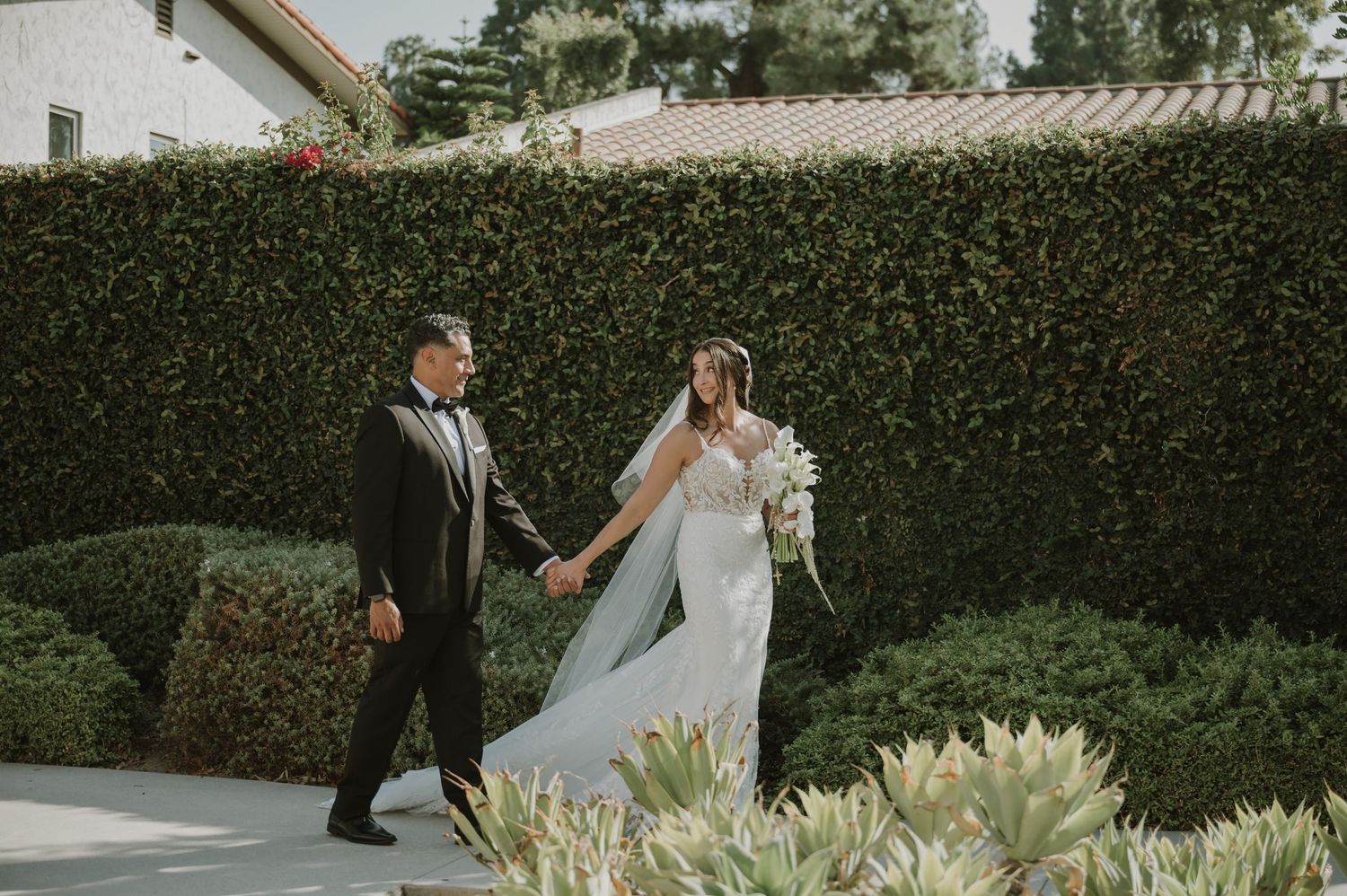 A bride and groom walk hand in hand through a lush green hedge garden on their wedding day.