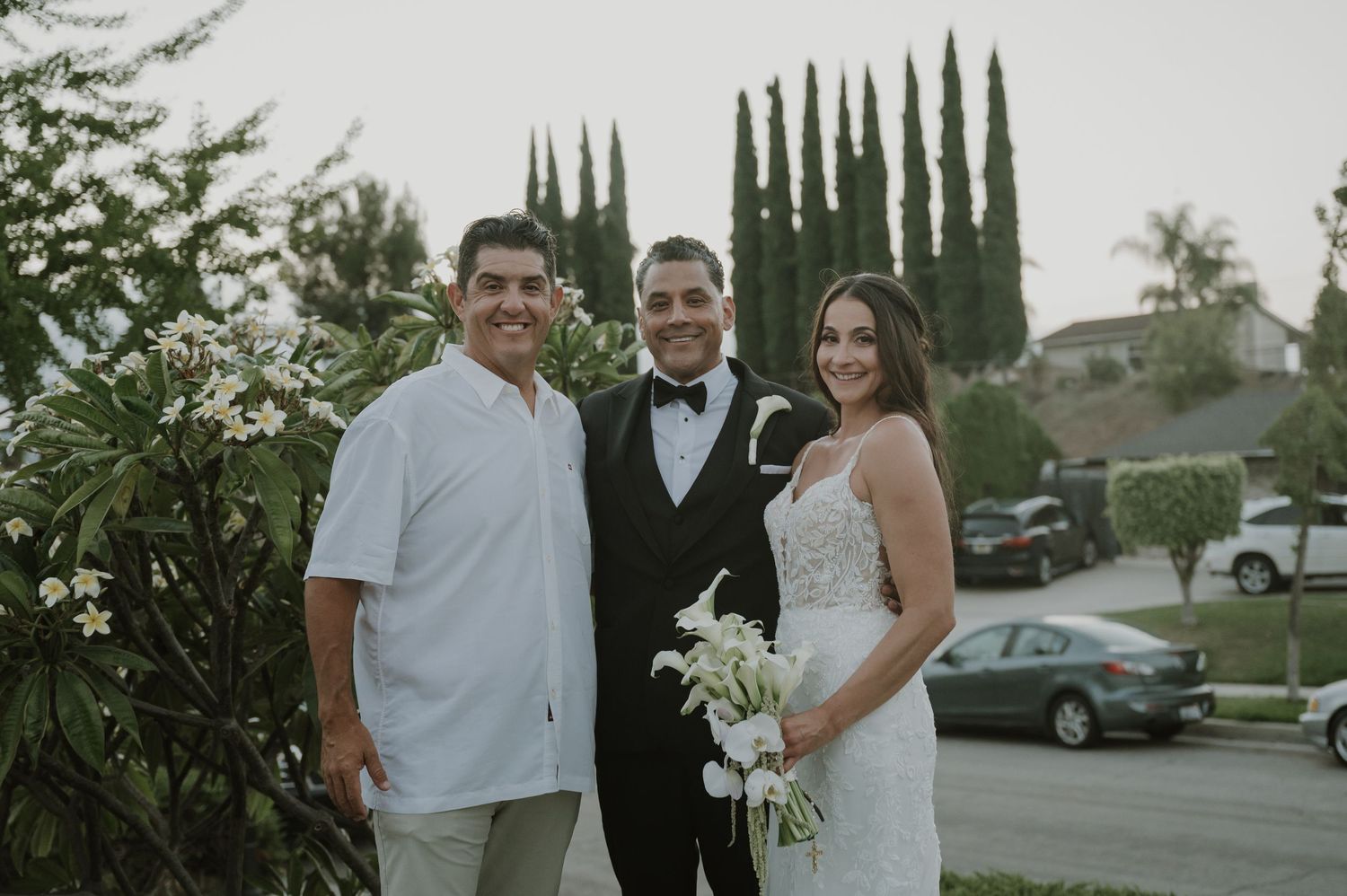A groom in a tuxedo poses with a bride holding white flowers and a guest in a white shirt outdoors.