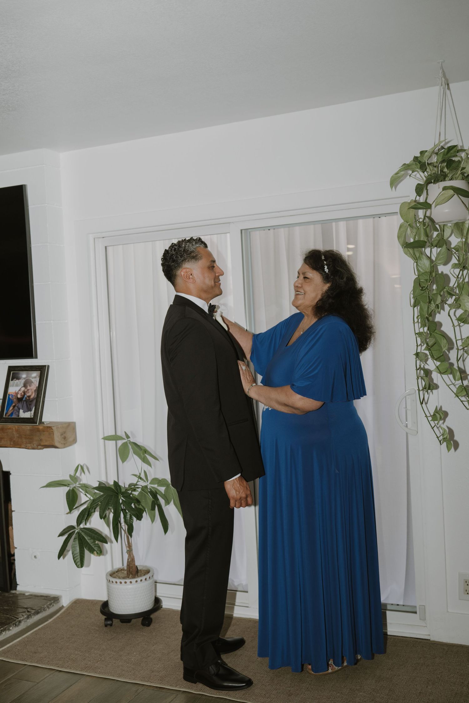 A woman in a blue dress adjusts a mans black tuxedo bow tie indoors before a wedding.