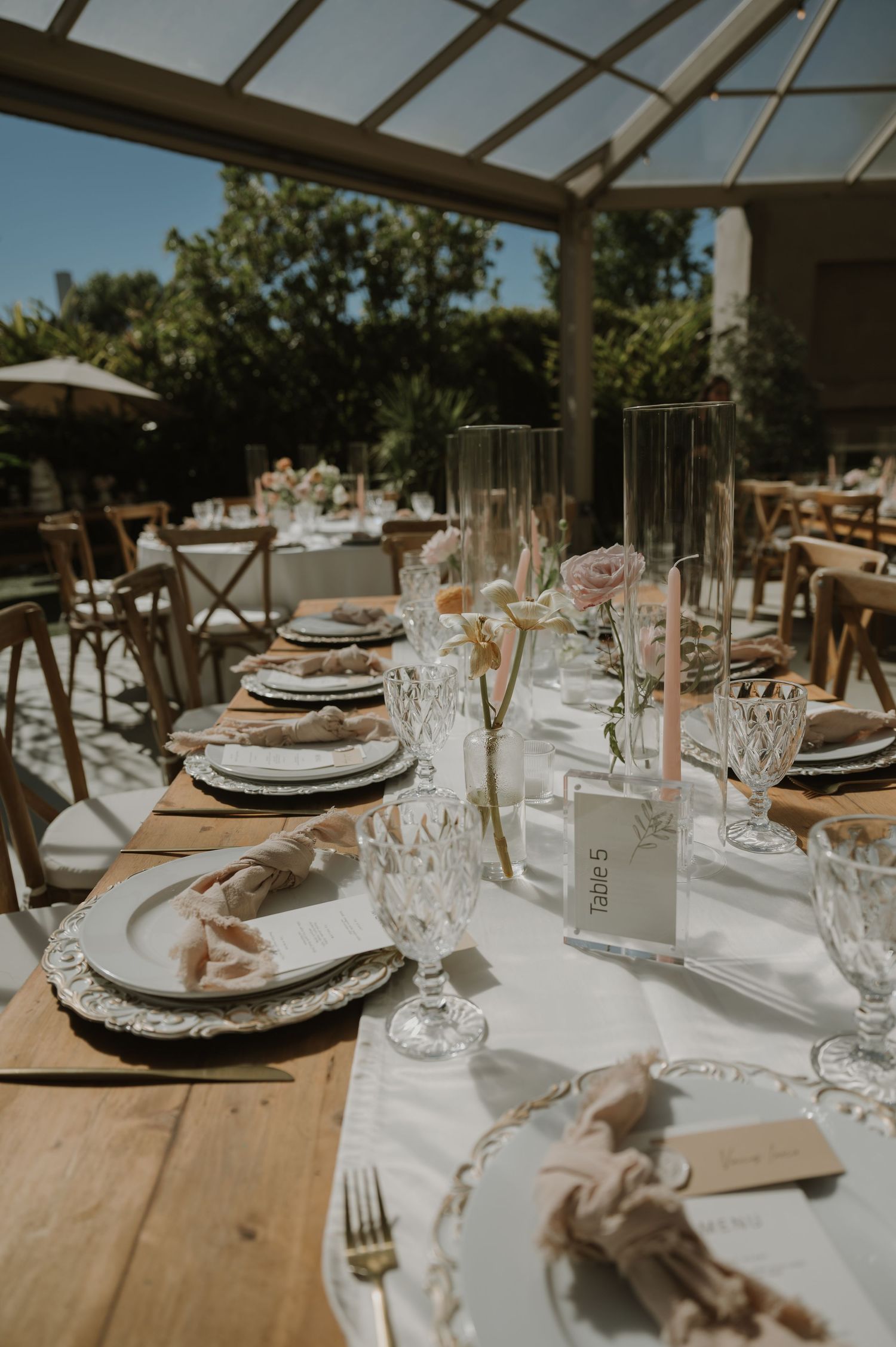 Elegant wedding table setting with crystal glassware, pink roses, white linens, and wooden chairs in a greenhouse.