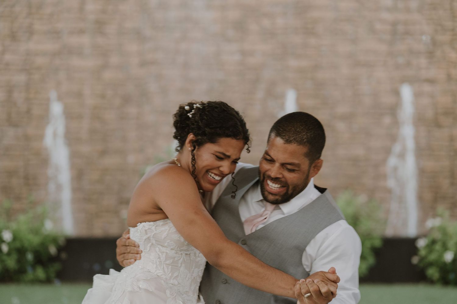 Joyful bride and groom laughing together during first dance in front of elegant fountain backdrop.
