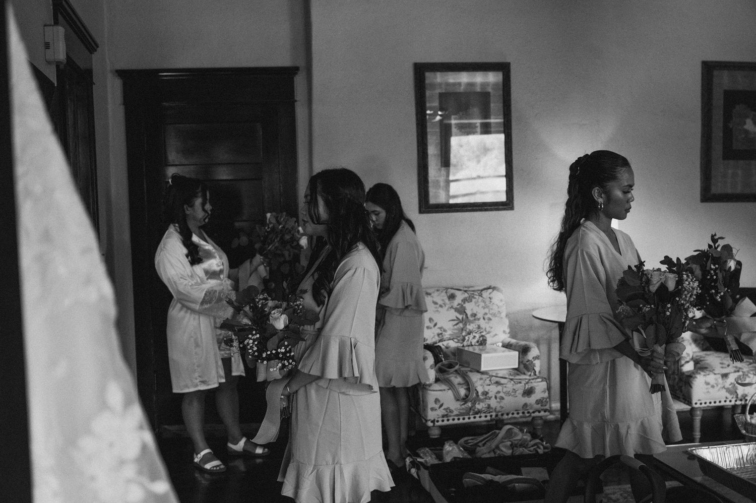 Bridesmaids in robes gather with floral arrangements in an elegant room during wedding preparations, captured in black and white.