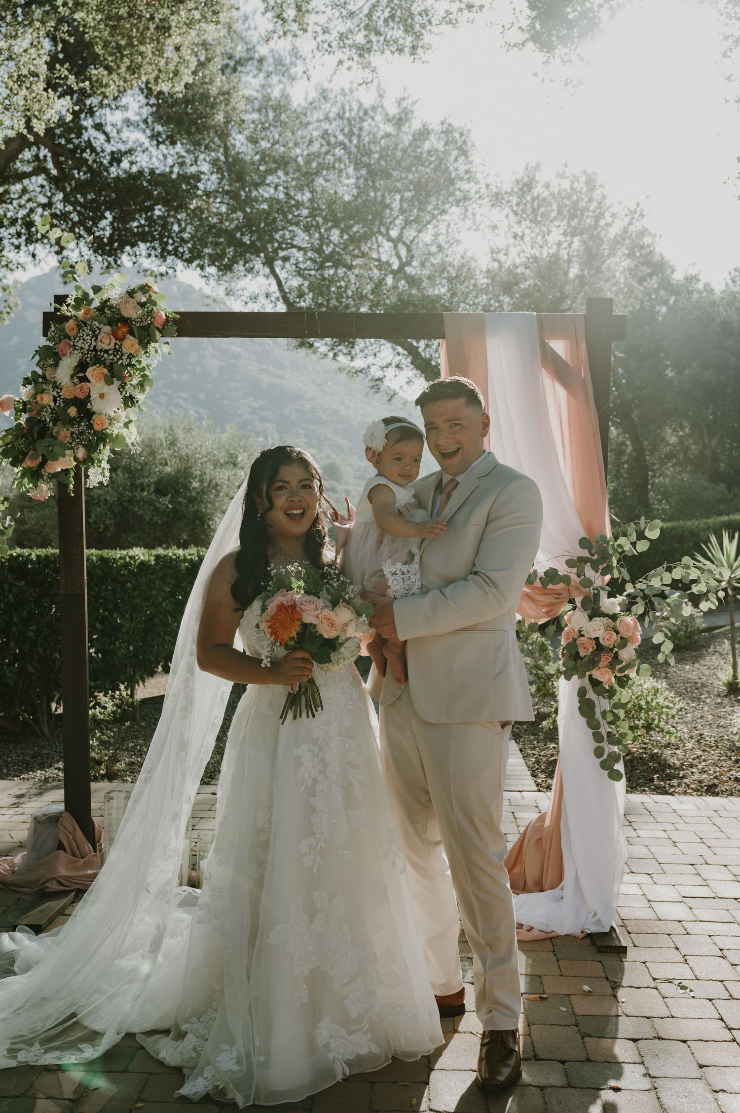 Bride and groom pose with their baby under a floral arch at an outdoor wedding ceremony.