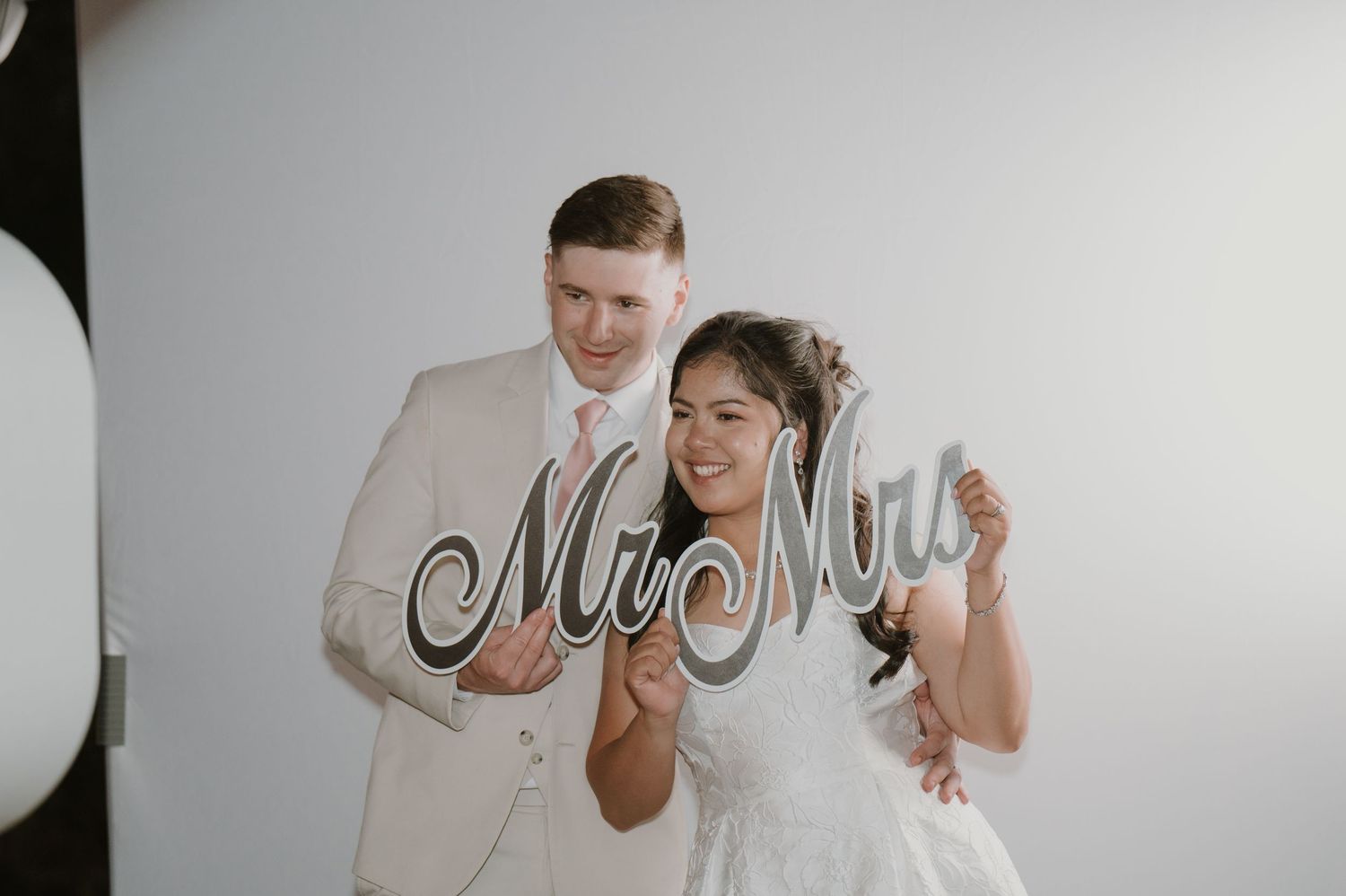 Smiling bride and groom pose with Mr and Mrs signs at wedding photo booth.