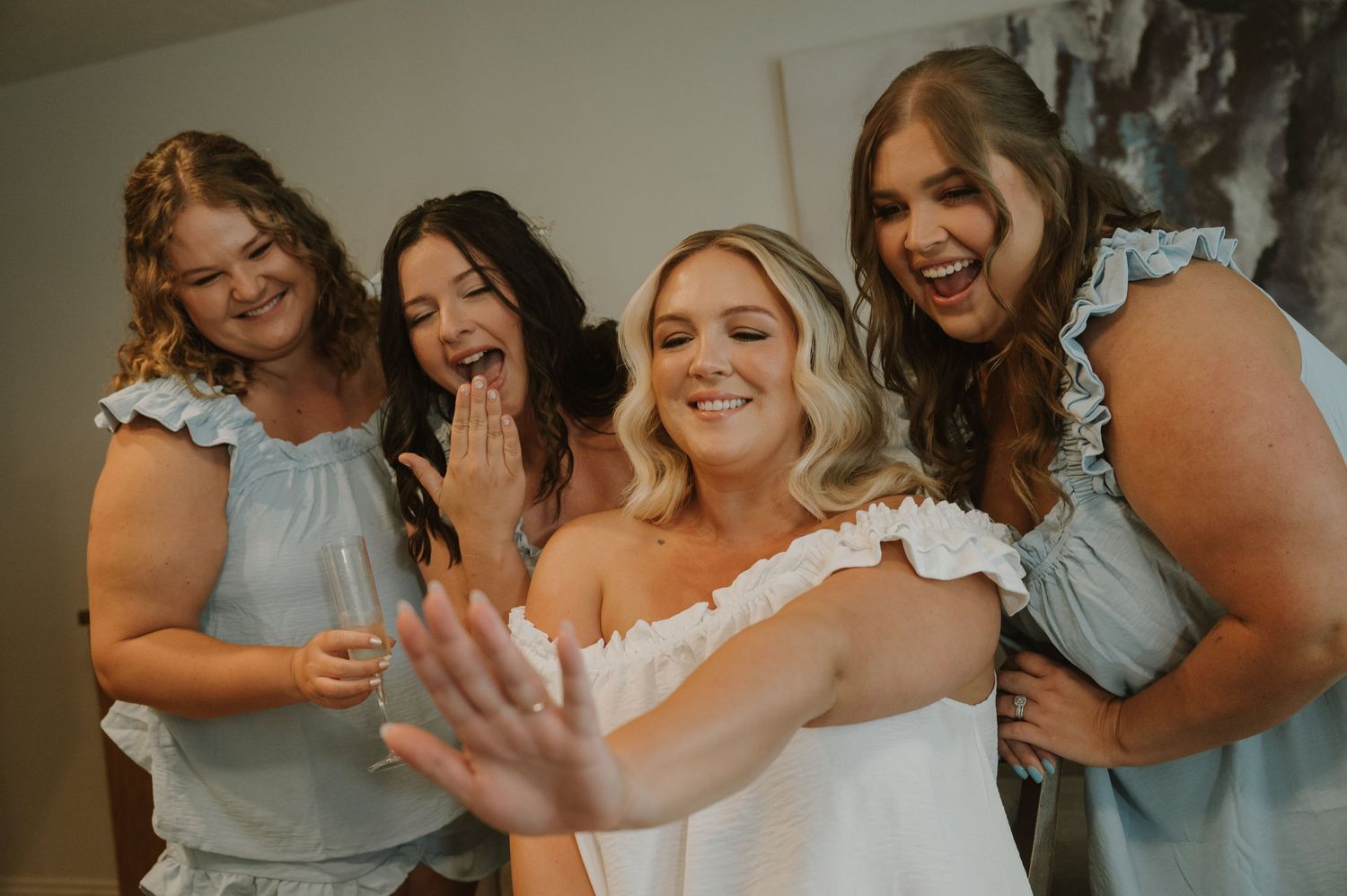 Bride in white dress laughs with three bridesmaids in light blue dresses during wedding preparations.