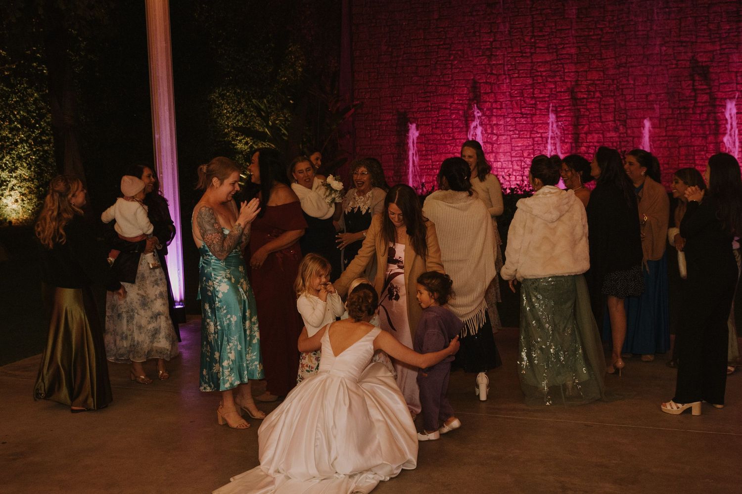 Wedding reception guests dancing outdoors at night with pink floral backdrop and bride in white gown.