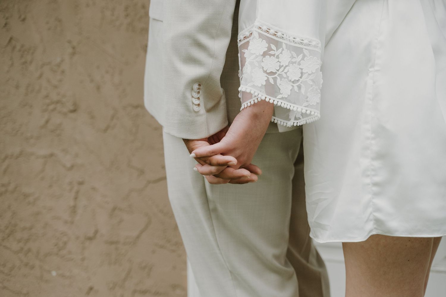 Couple holding hands in white wedding attire, showcasing elegant lace sleeve detail against a neutral textured wall.