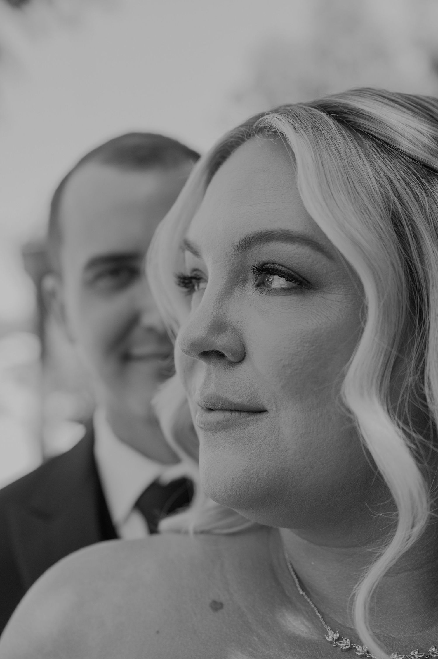 Black and white wedding photo of a smiling groom and elegant bride with flowing blonde hair gazing thoughtfully.