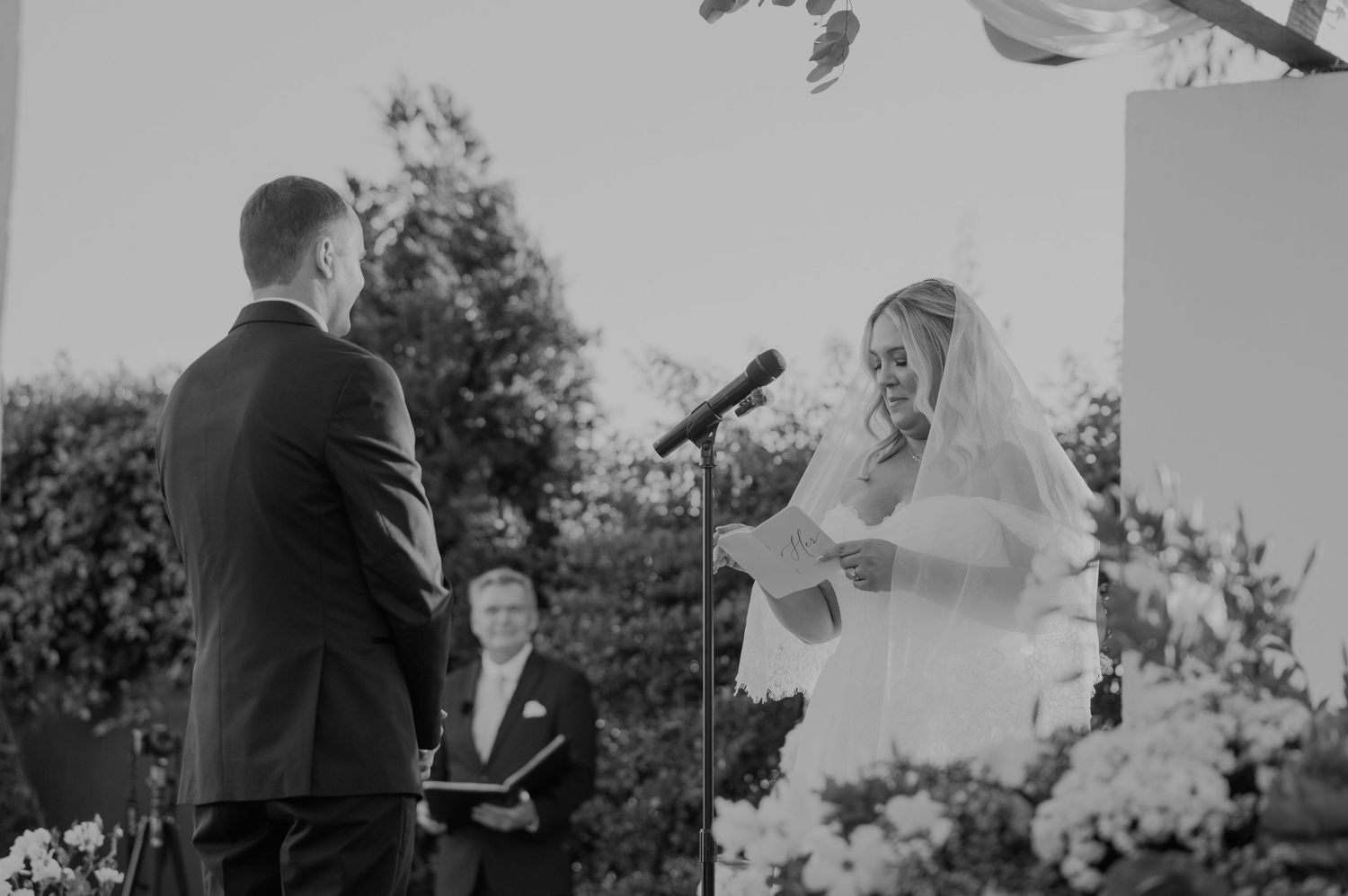 Black and white photo of bride reading vows to groom at outdoor wedding ceremony altar.