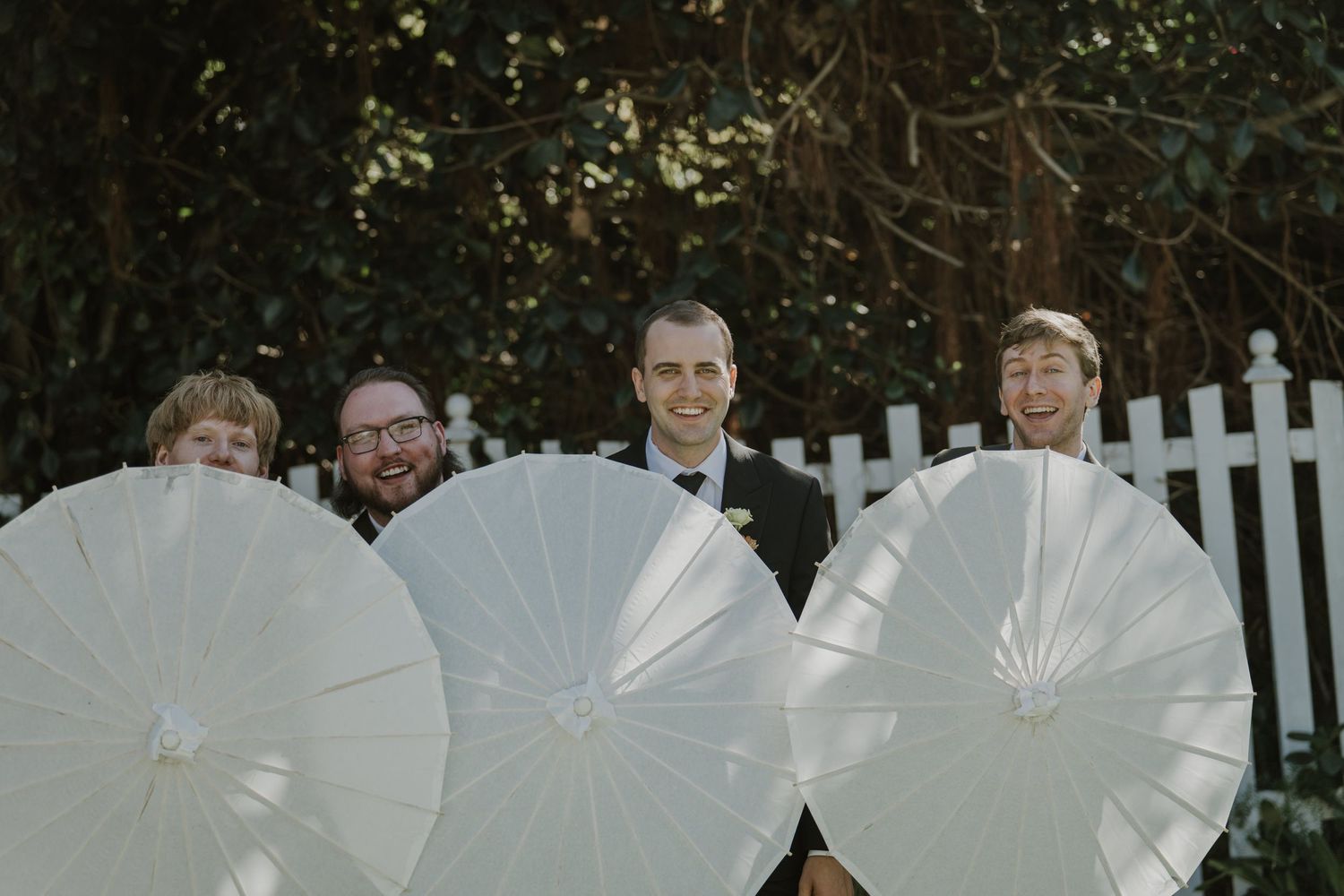 Wedding party smiling behind white paper parasols outdoors near white picket fence.