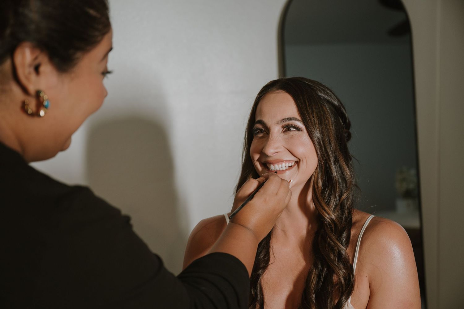 A makeup artist carefully applies finishing touches to a smiling bride during wedding preparations.