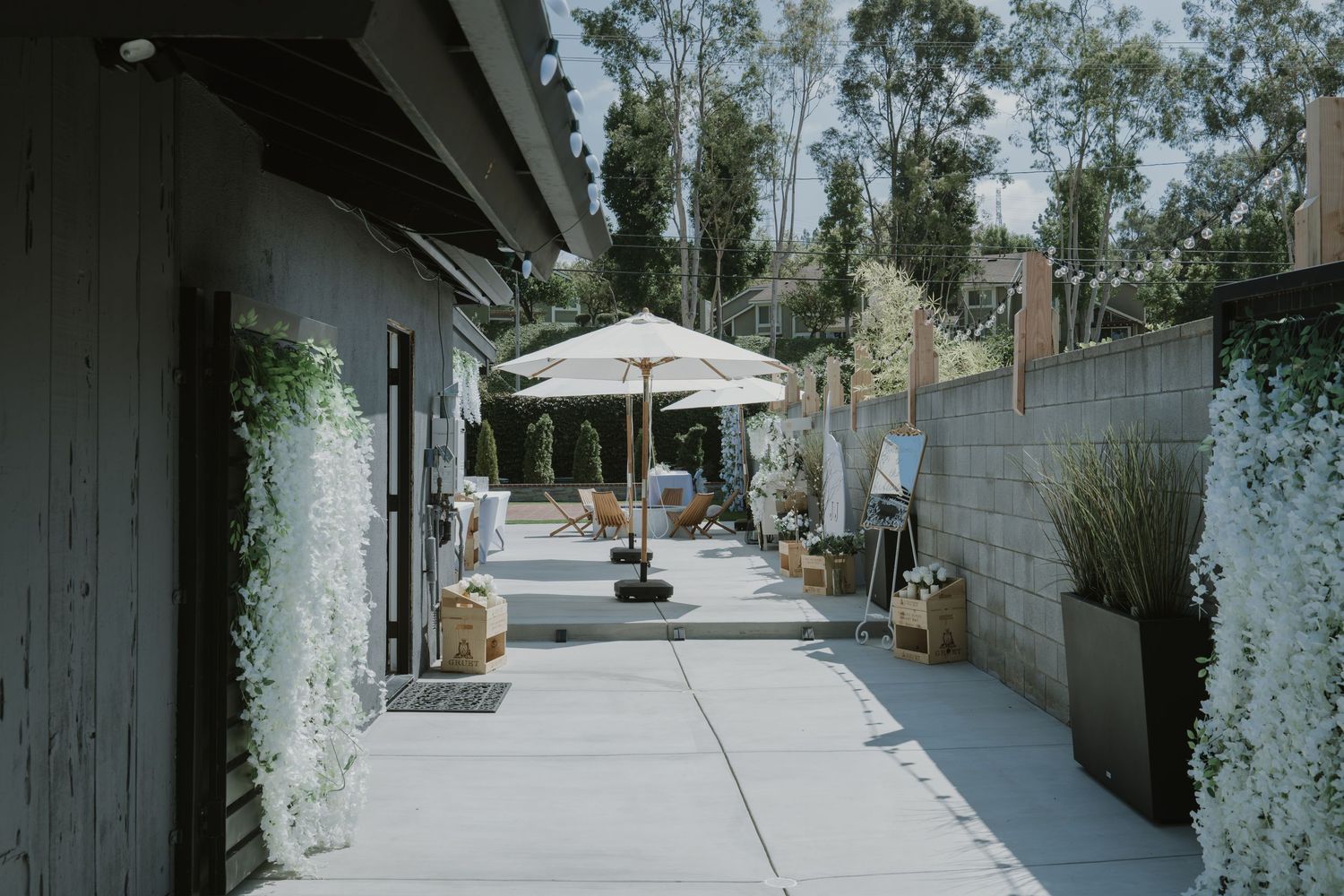 Serene outdoor wedding venue pathway with white floral walls, umbrella, and lush greenery.